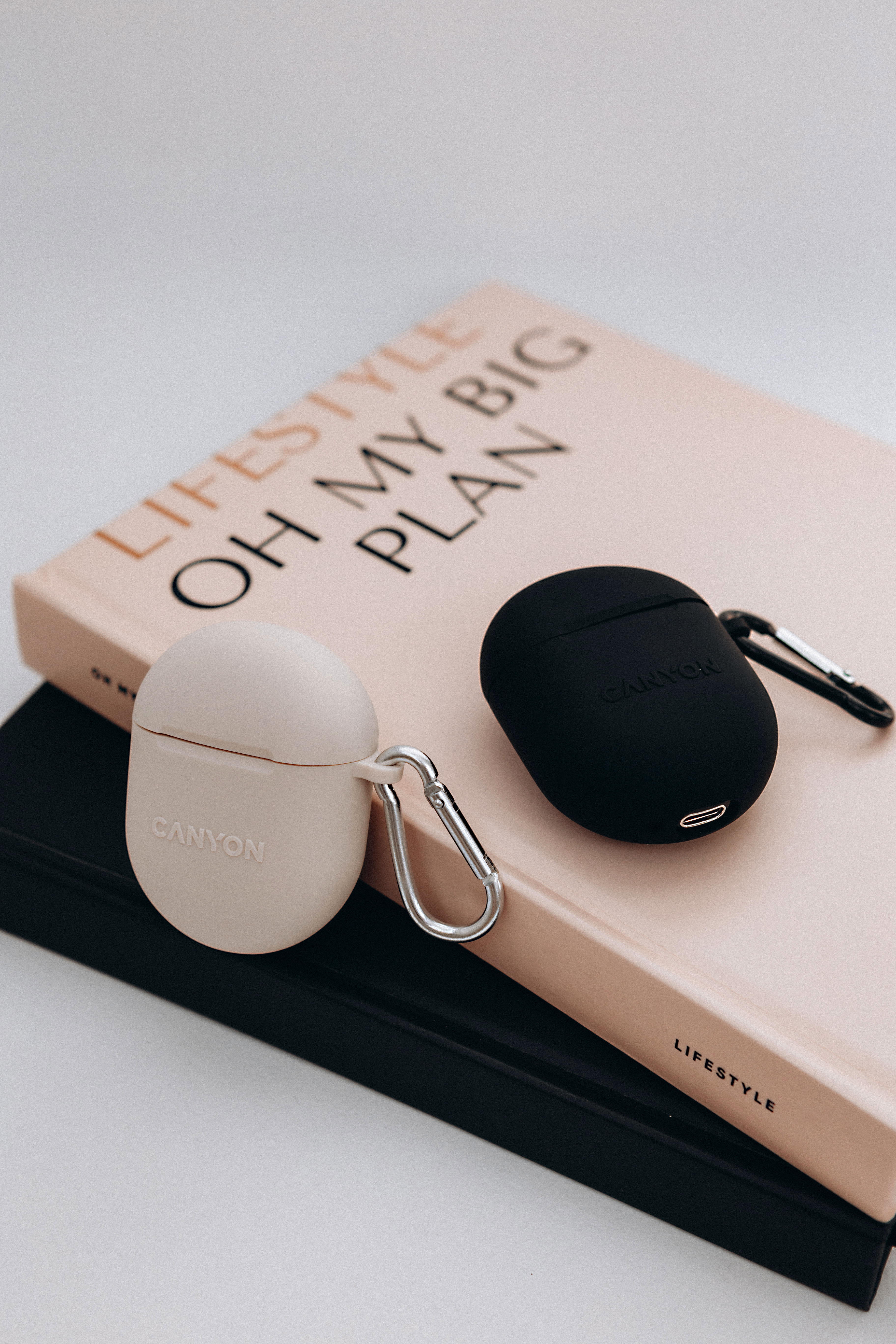 Dwa etui na słuchawki Canyon, jedno beżowe, jedno czarne, na książkach.