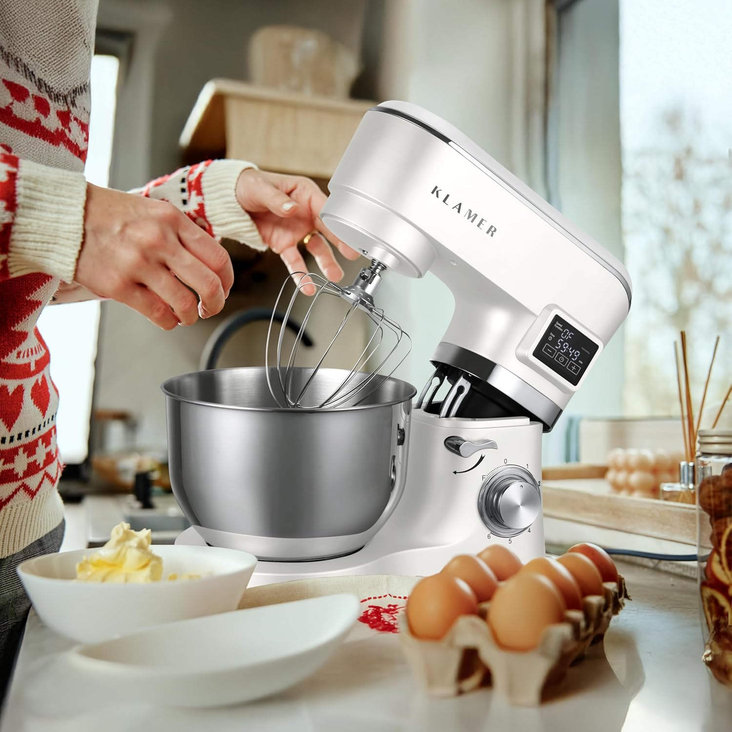 Eine weiße Küchenmaschine mit Rührbesen, Schüssel, Eiern und Butter auf einem Tisch.