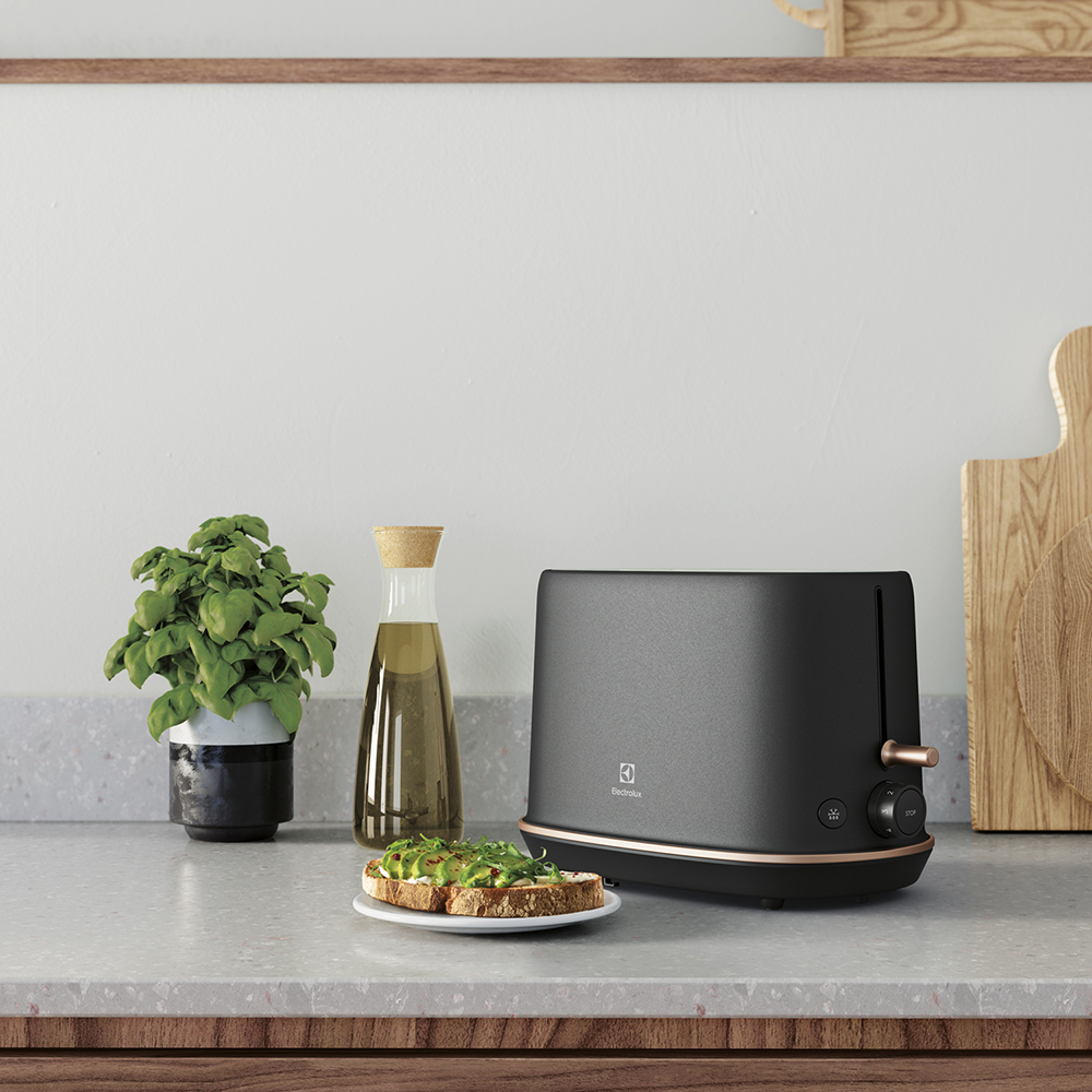 Czarny toster z akcentami w kolorze różowego złota, obok tostów z awokado i butelki oleju.