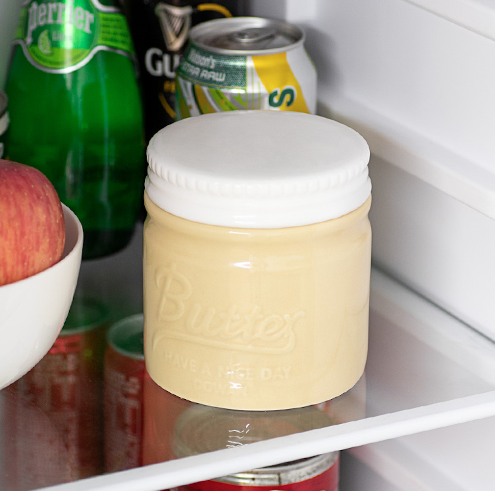 Eine Butterdose mit weißem Deckel steht im Kühlschrank neben Getränken und einem Apfel.
