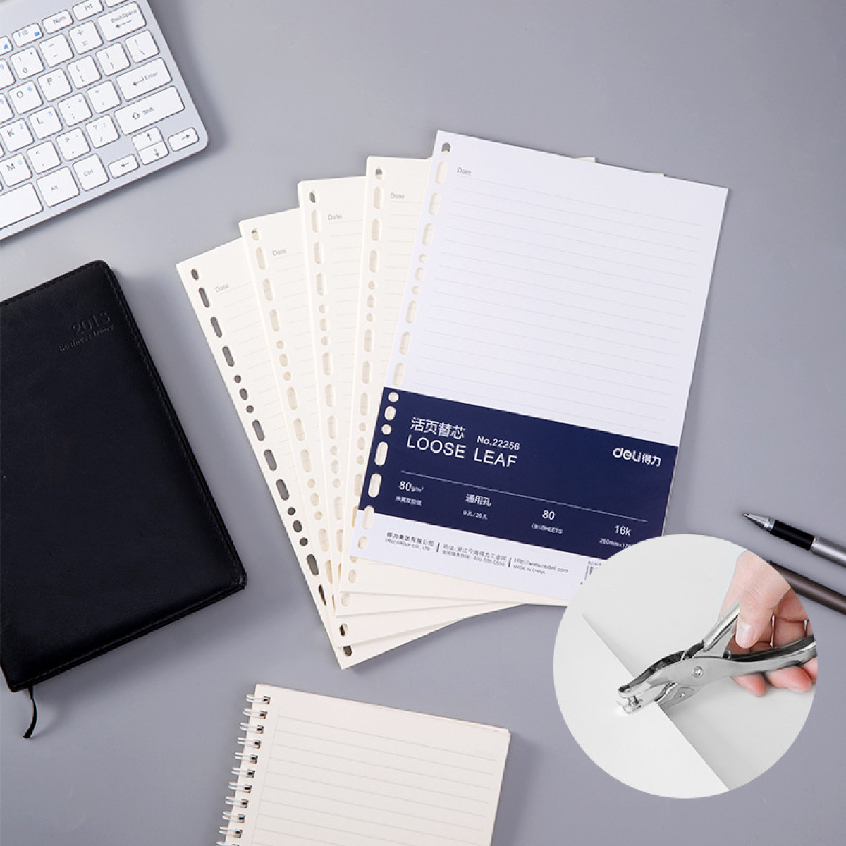 Scena biurkowa z notatnikiem, klawiaturą, długopisami, luźnymi kartkami i dziurkaczem.