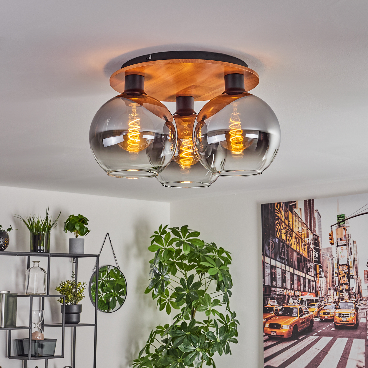 Deckenleuchte mit drei Rauchglas-Kugeln und Holzsockel, leuchtet über einem Regal mit Pflanzen.