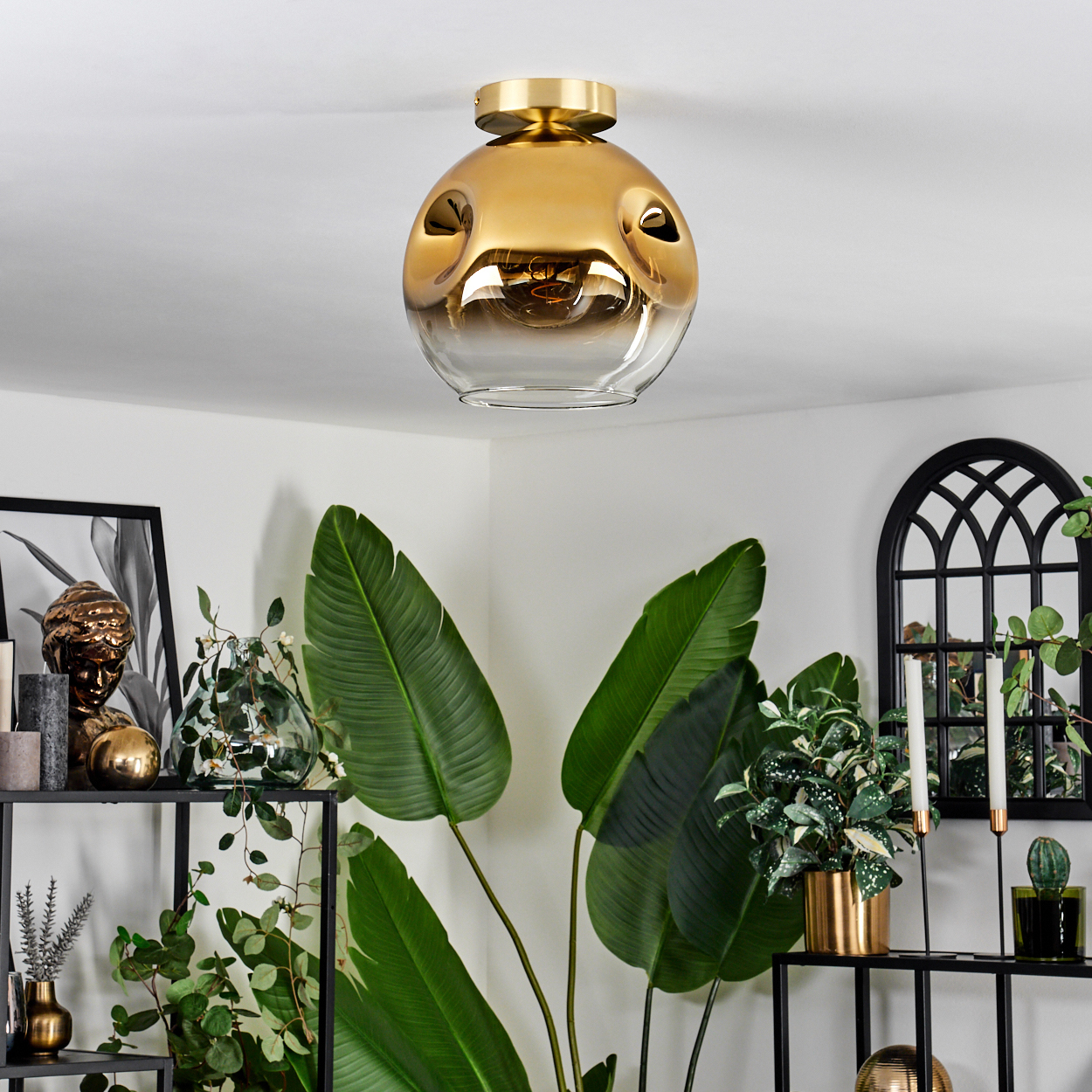 Eine Deckenleuchte aus Gold und Glas hängt über einem Regal mit Pflanzen und Deko.