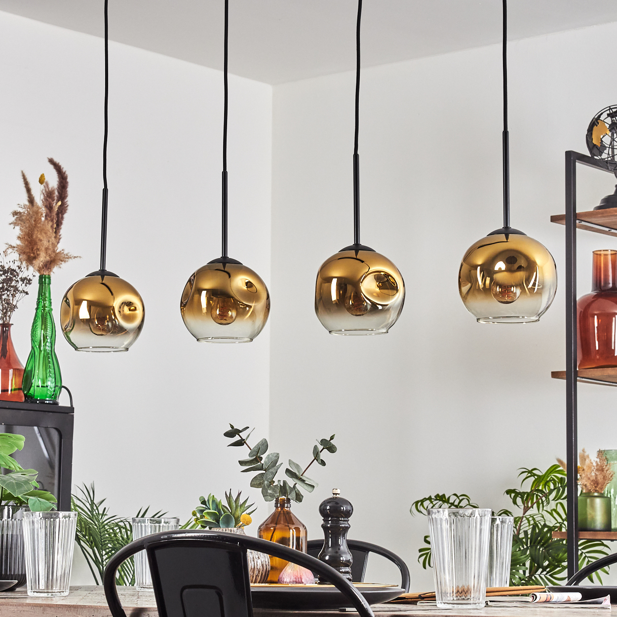 Vier goldene Pendelleuchten über einem Tisch mit Gläsern und Pflanzen.