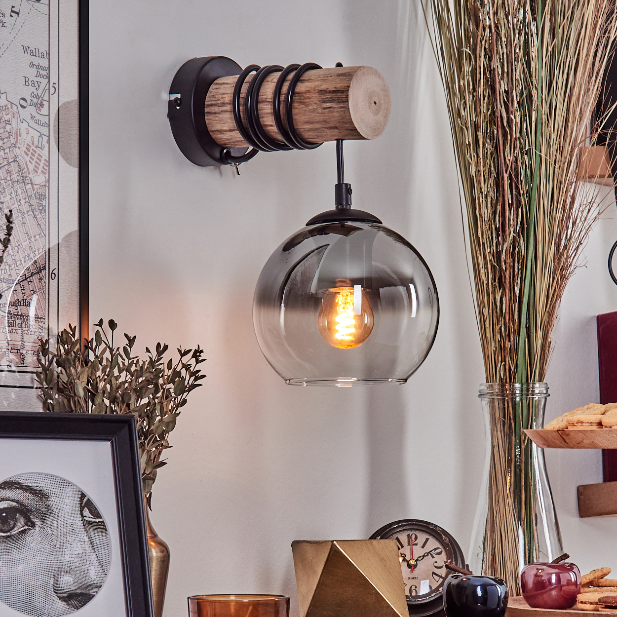 Eine Wandleuchte mit Holzarm und Rauchglas-Schirm hängt über Dekorationen.