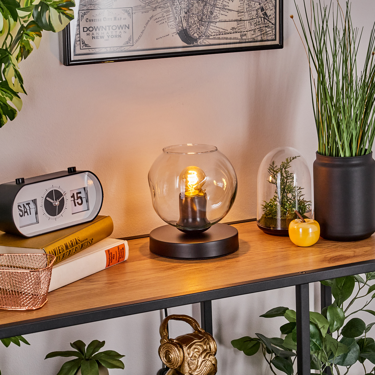 Ein Schreibtisch mit einem Flip-Clock, Büchern, einer Glaslampe und Pflanzen. Eine Karte hängt an der Wand.