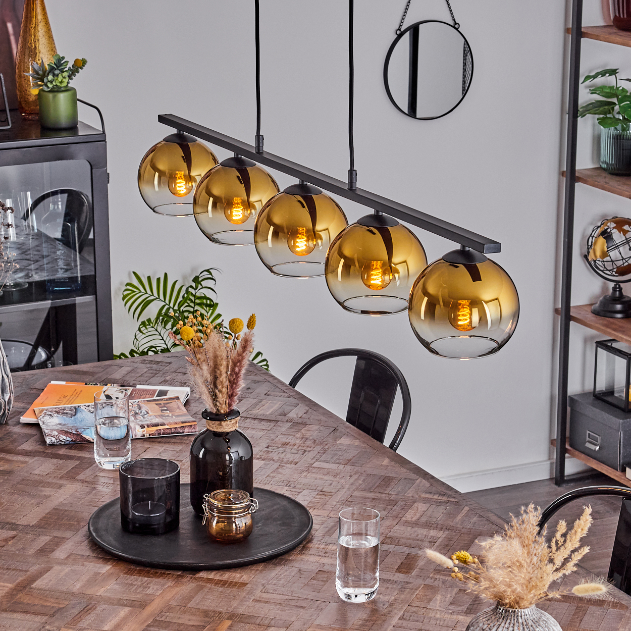 Hängelampe mit bernsteinfarbenen Glaskugeln über einem Holztisch mit Vasen und Gläsern.
