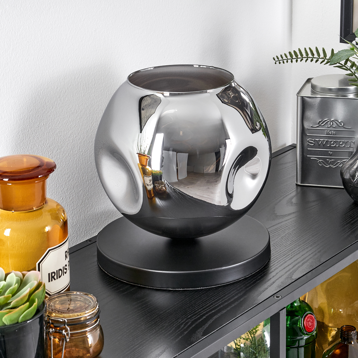 Eine silberne, kugelförmige Lampe mit unregelmäßiger Oberfläche steht auf einem schwarzen Sockel.