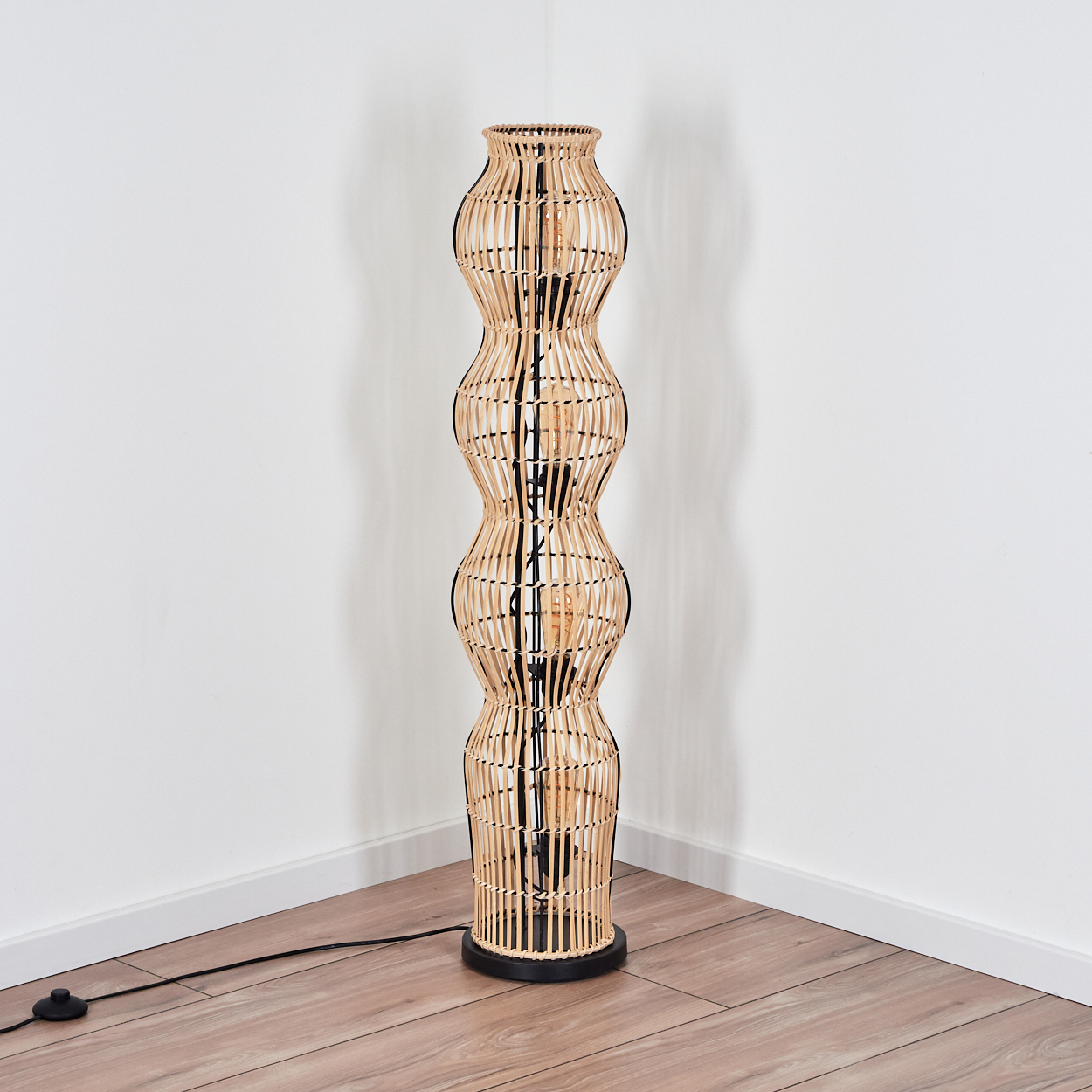 Ein hoher Stehleuchte aus hellem Rattan mit geschwungenem Design steht in einer Ecke.