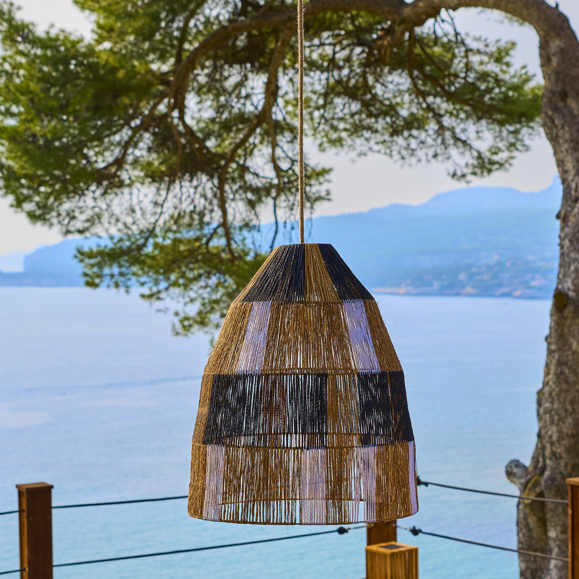 Ein geflochtener Lampenschirm hängt vor dem Meer, mit einem Baum und Bergen im Hintergrund.