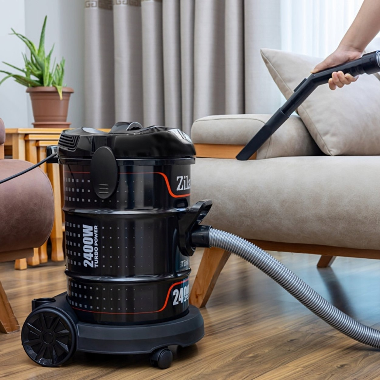 Czarny odkurzacz czyści beżową sofę wężem.