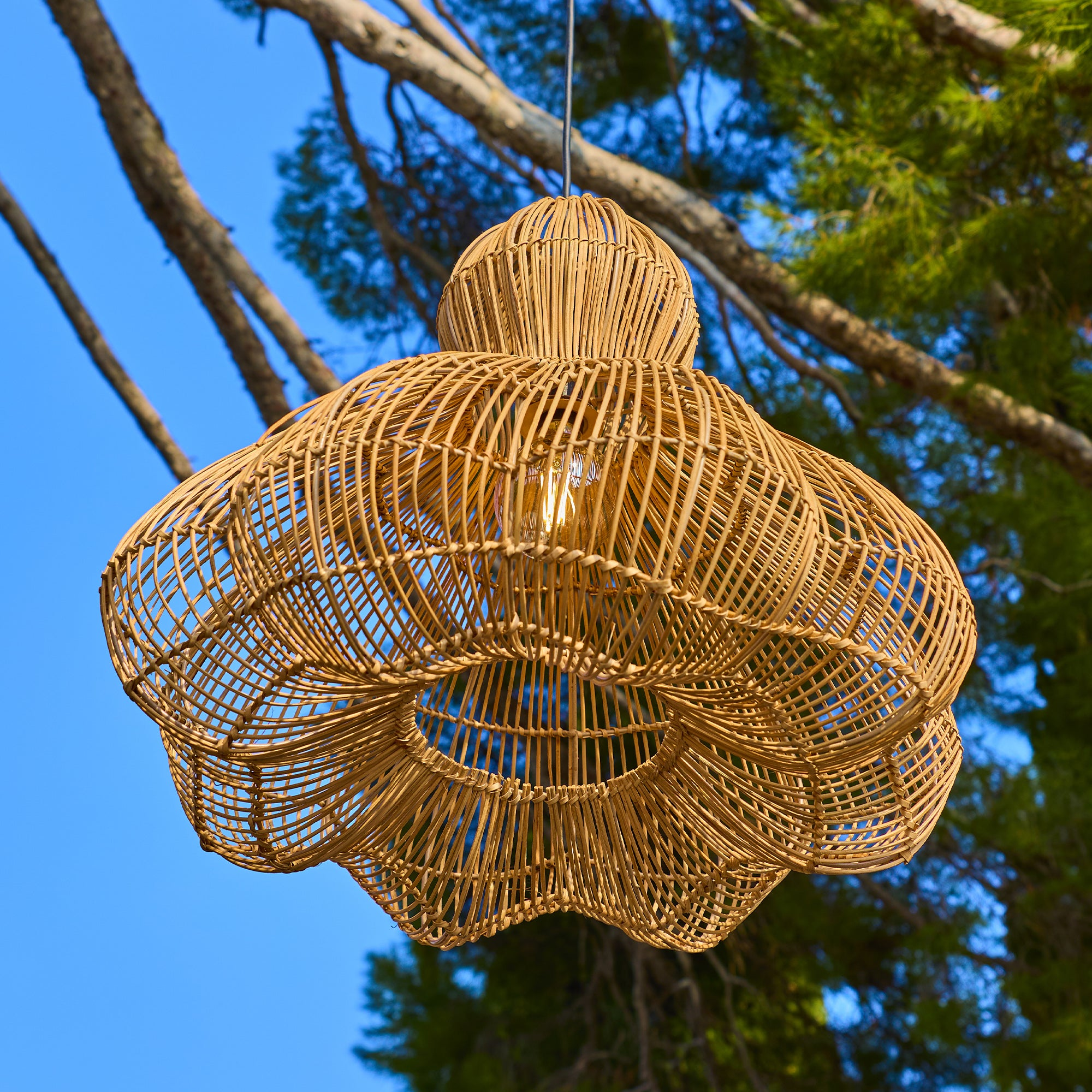 Ein geflochtener Lampenschirm aus Rattan hängt vor blauem Himmel und Bäumen.