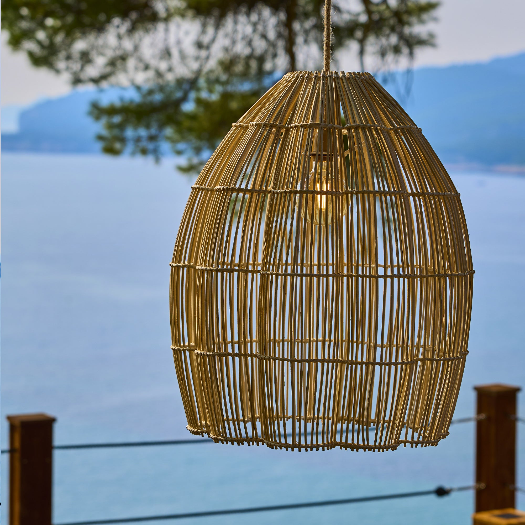 Ein runder, geflochtener Lampenschirm aus hellem Holz hängt draußen mit Meerblick.