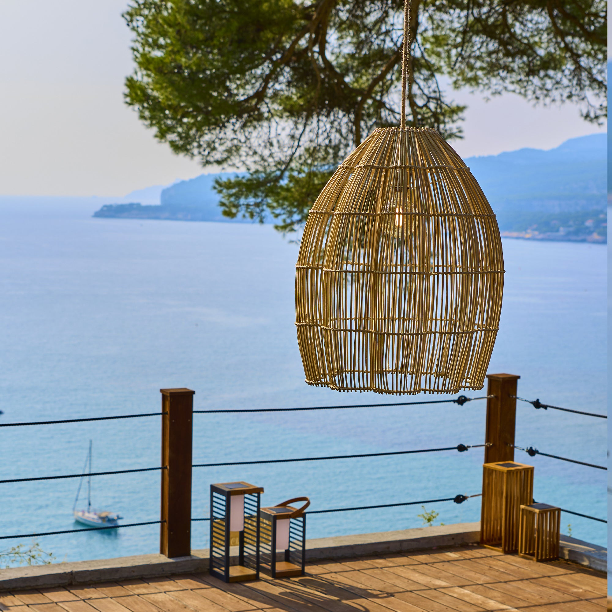 Ein Weidenkorb-Hängelampenschirm über einem hölzernen Deck mit Blick aufs Meer.