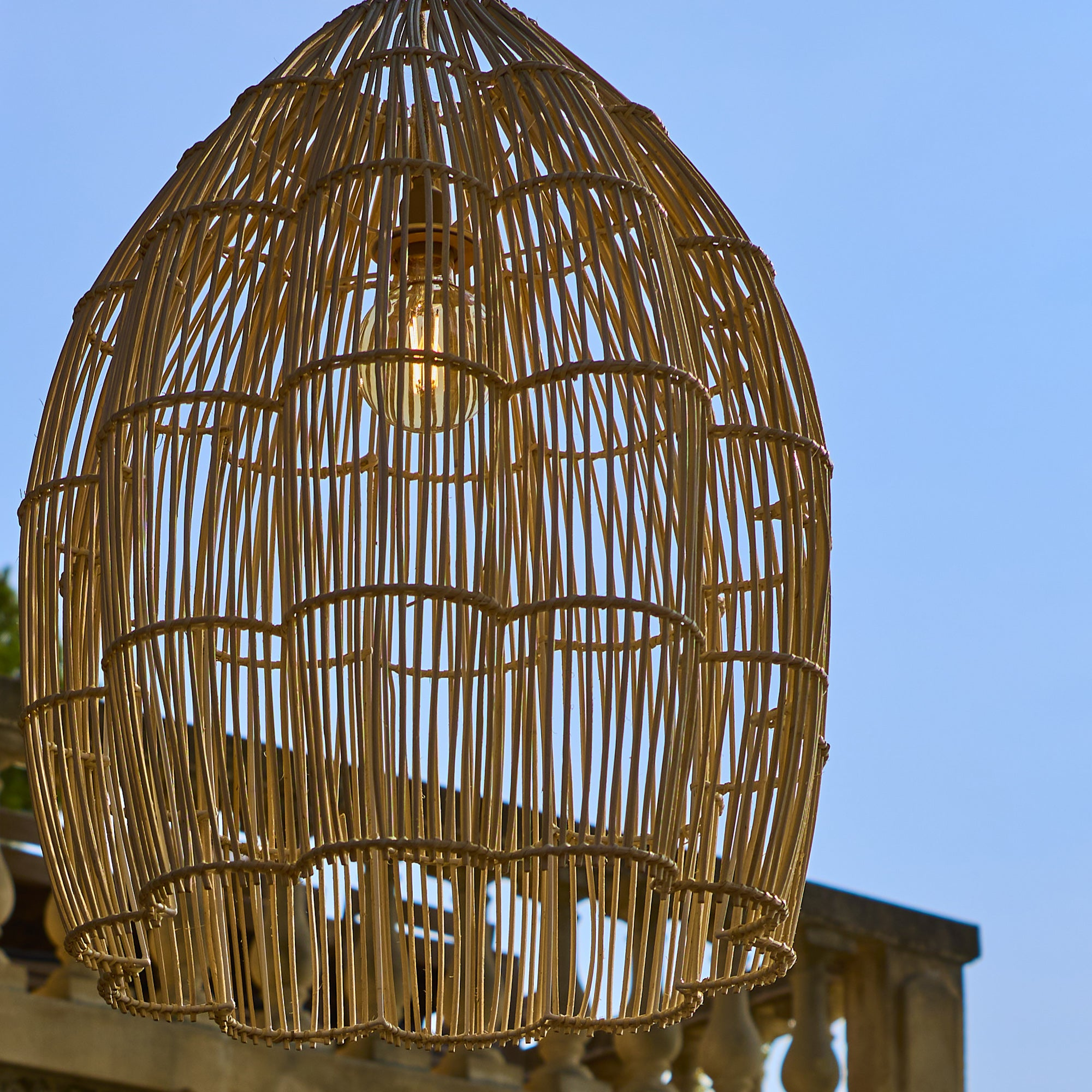 Eine Korblampe aus Rattan hängt vor blauem Himmel mit einer Glühbirne.