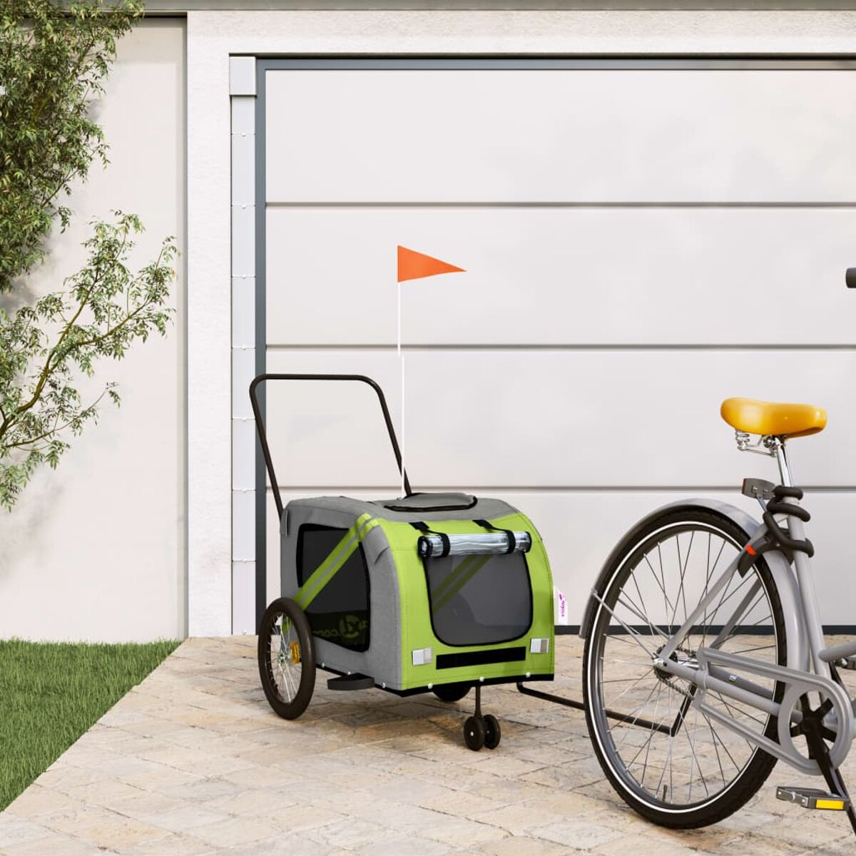 Ein grüner Fahrradanhänger mit orangefarbener Fahne steht neben einem Fahrrad vor einer Garage.
