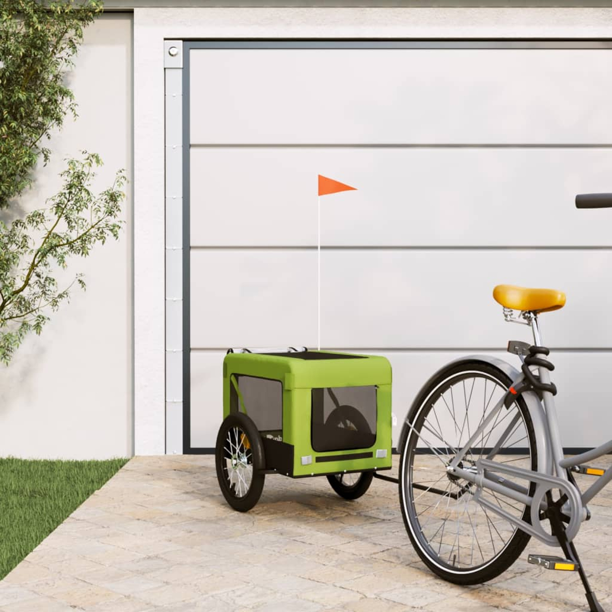 Ein grüner Fahrradanhänger mit orangefarbener Fahne steht neben einem Fahrrad vor einer Garage.
