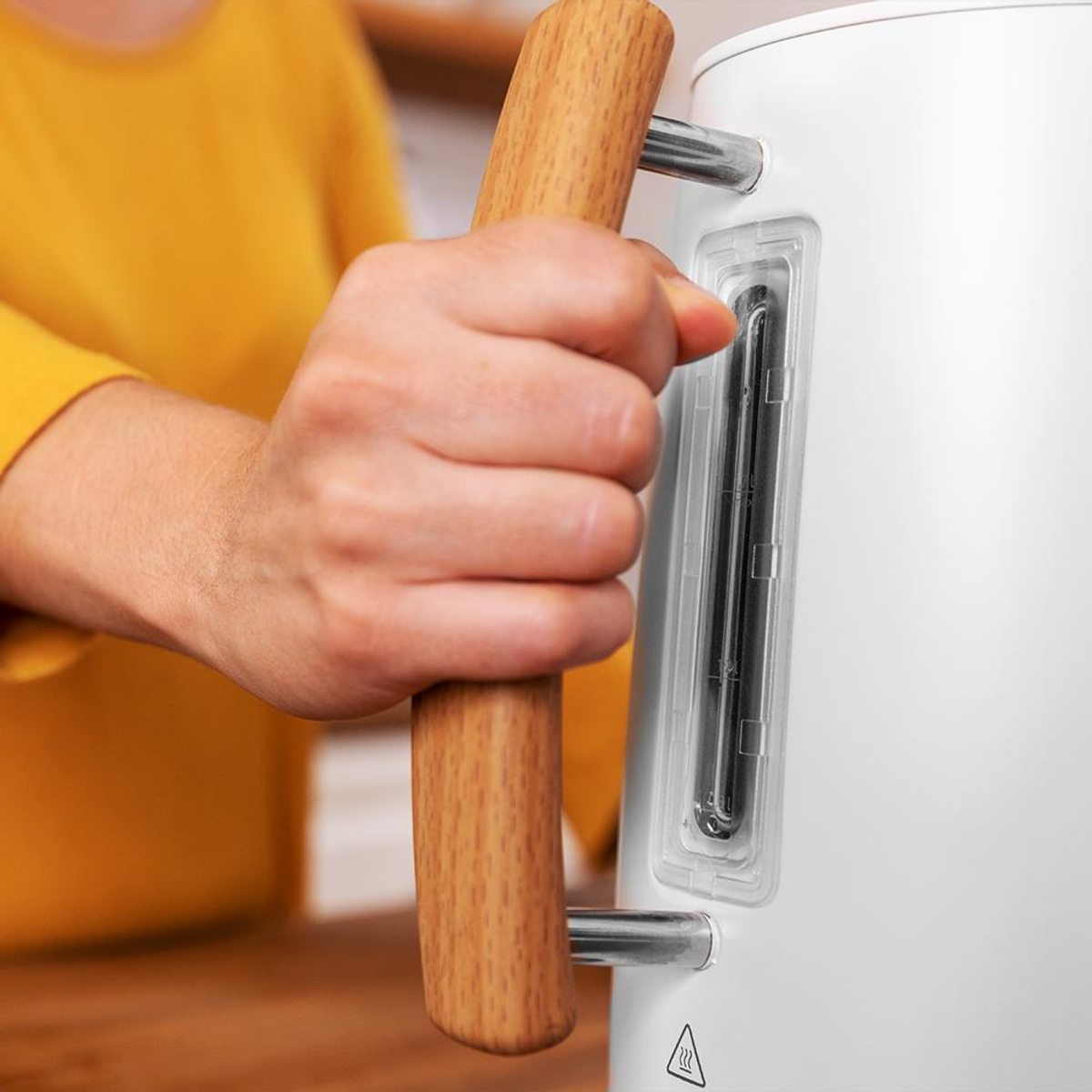 Dłoń w żółtym swetrze chwyta drewnianą rączkę białego czajnika.