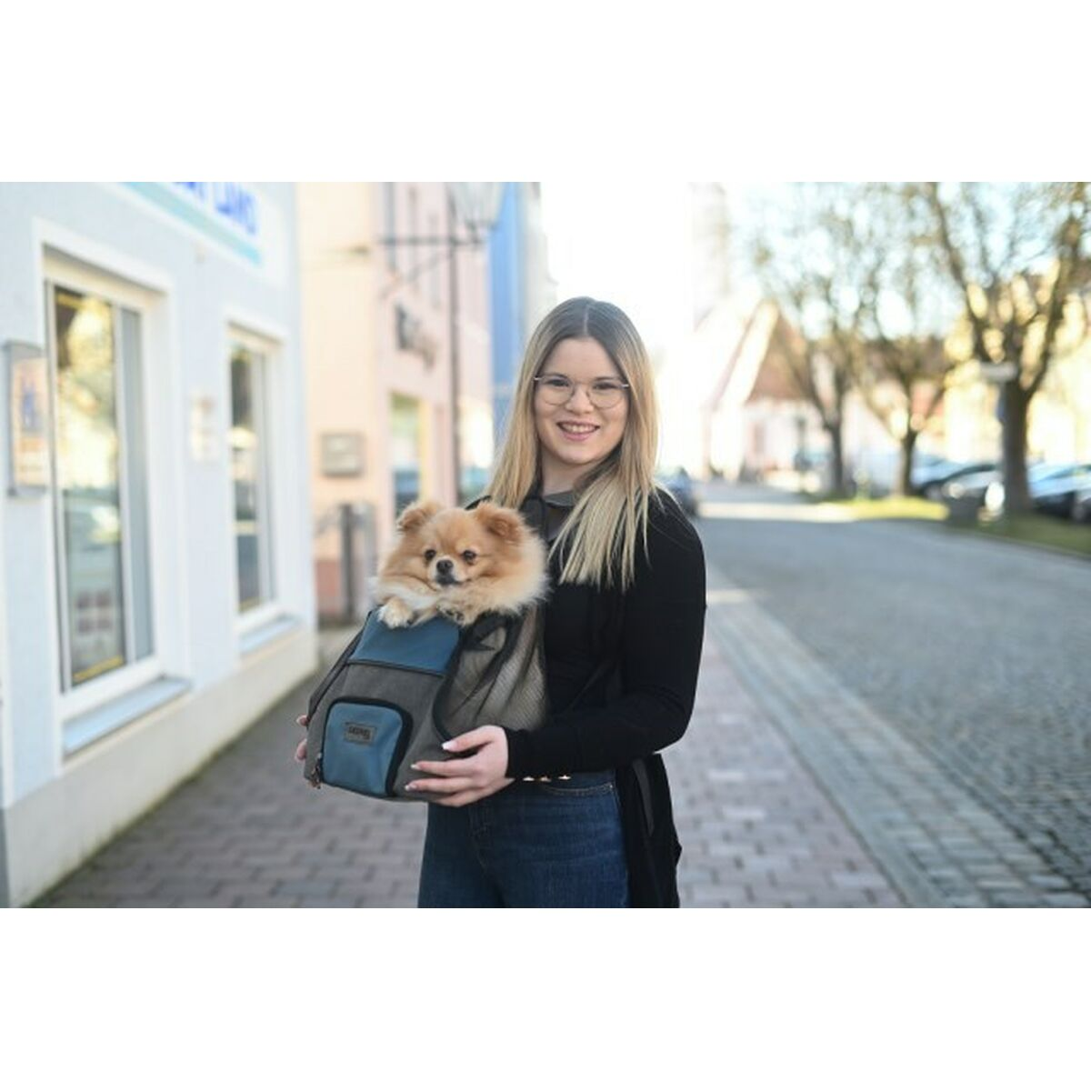 Una mujer con gafas lleva un perro Pomerania pequeño en una bolsa de transporte azul y gris en una calle.