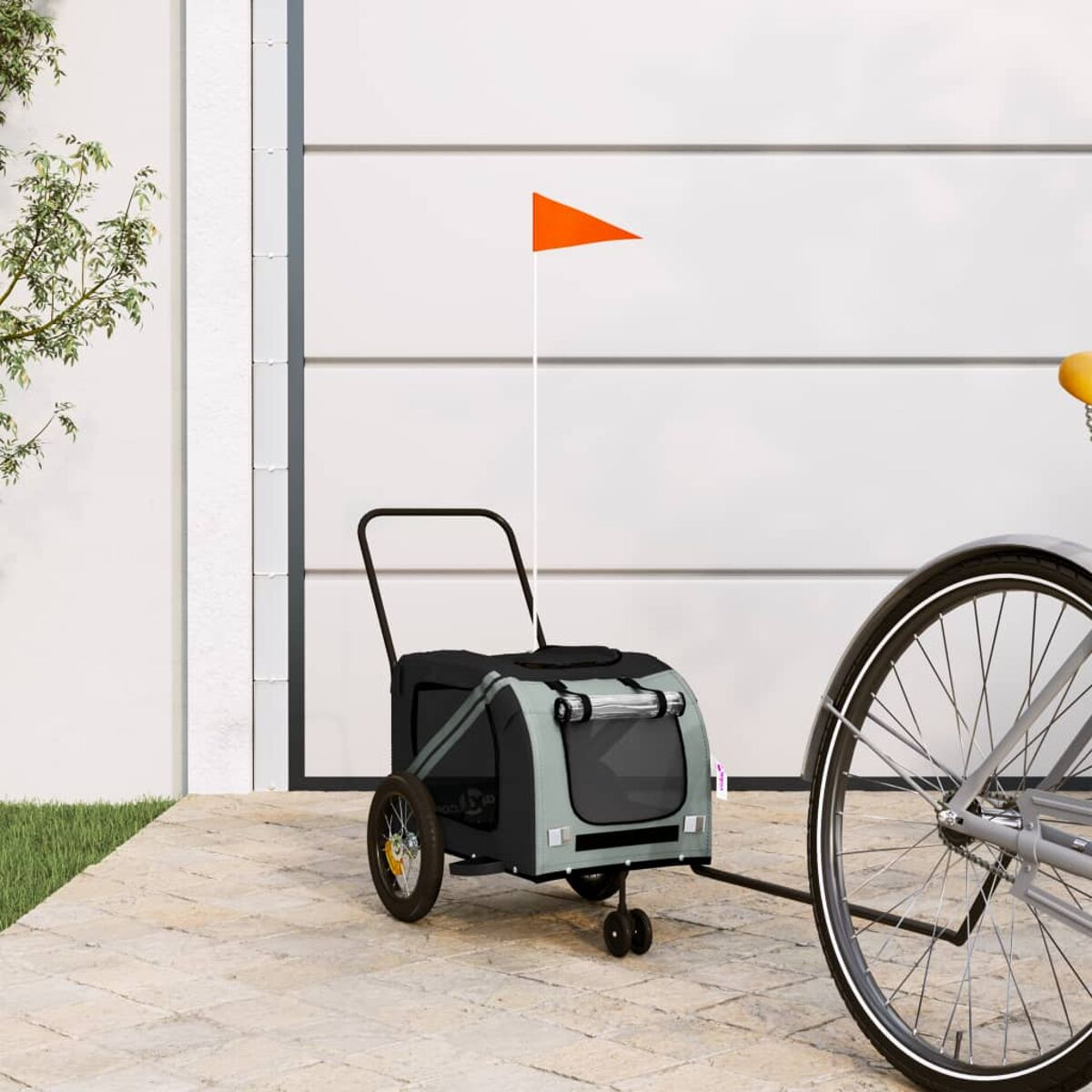 Ein grauer Fahrradanhänger mit orangefarbener Flagge steht auf einem Gehweg vor einer Garagentür.