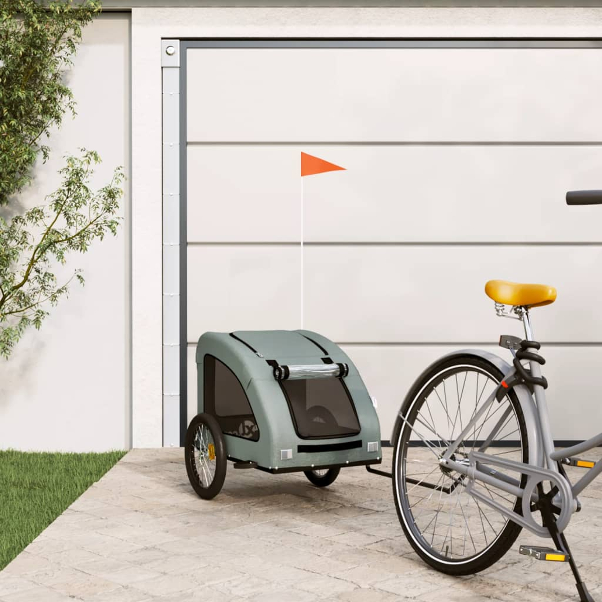 Ein grauer Fahrradanhänger mit orangefarbener Flagge steht neben einem Fahrrad vor einer Garage.