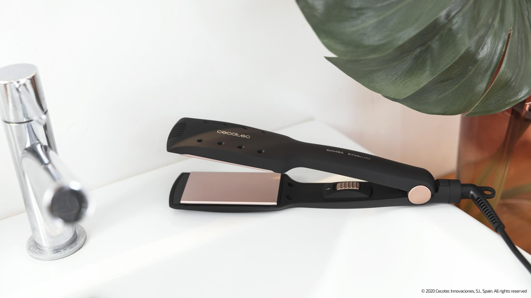 Una piastra per capelli nera con piastre color oro rosa poggia su un lavandino bianco vicino a un rubinetto.