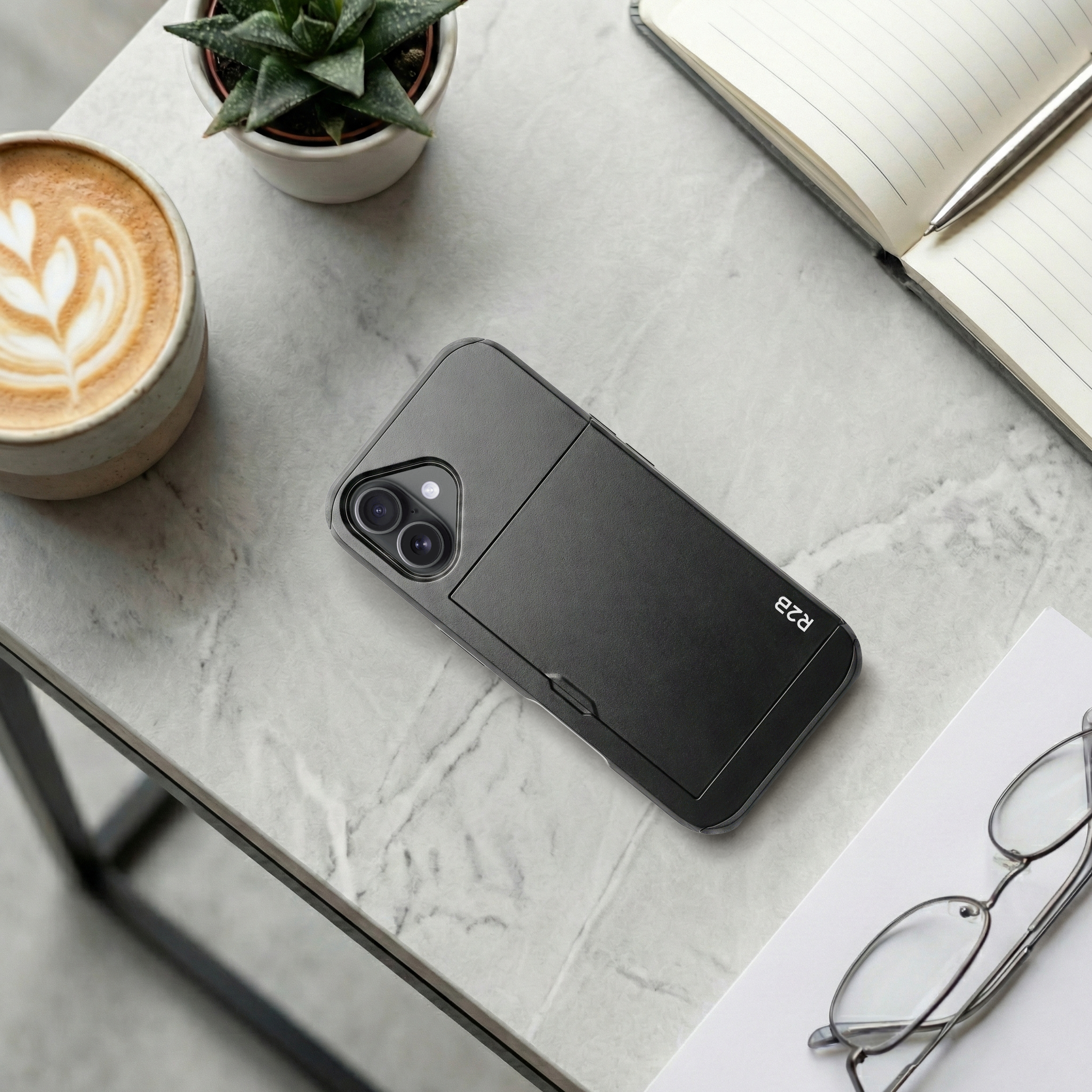 Zwarte telefoon met cameralenzen naast koffie, plant, notitieboek en bril.