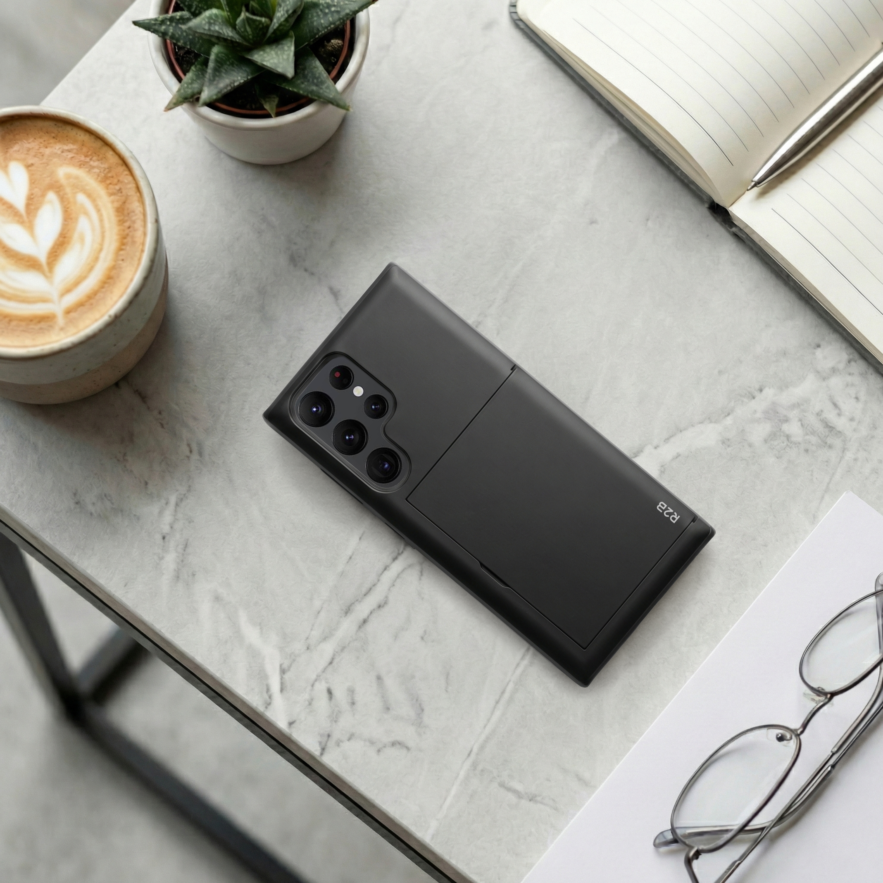 Ein schwarzes Smartphone liegt auf einem Marmortisch neben Kaffee, Pflanze und Notizbuch.