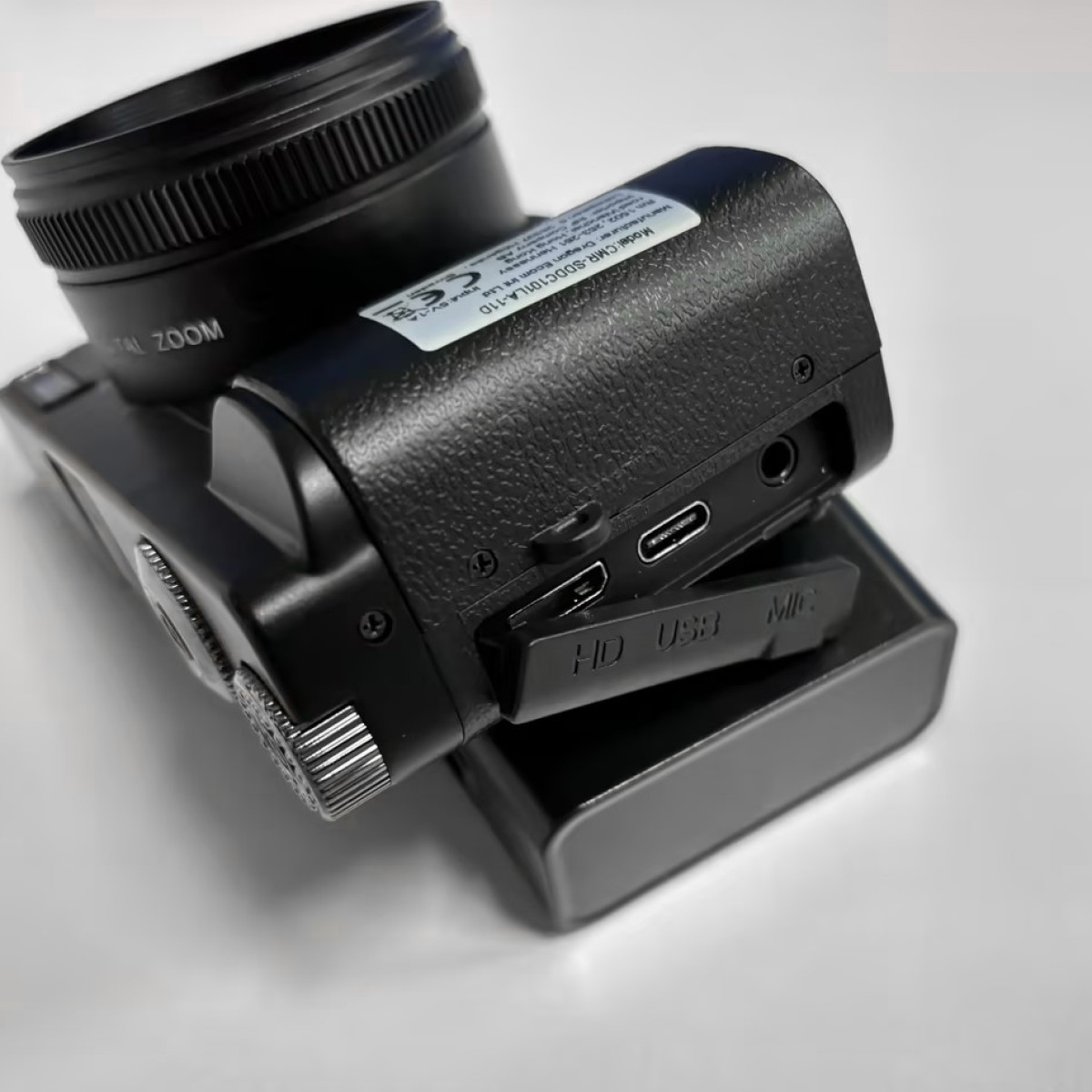 Czarny aparat cyfrowy z obiektywem, portami i baterią na białym tle.