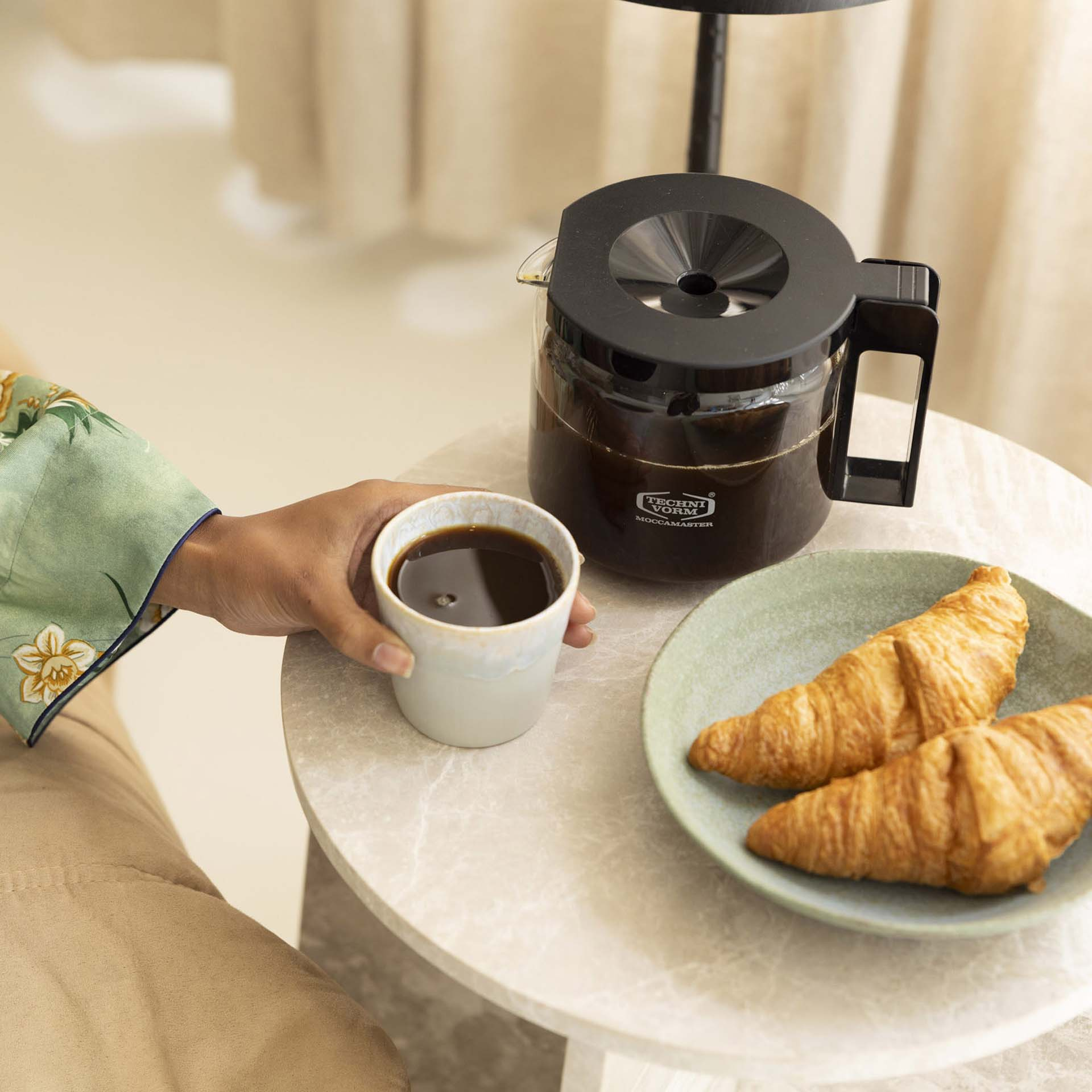 Une main tient une tasse de café à côté d'une cafetière et de croissants sur une table.
