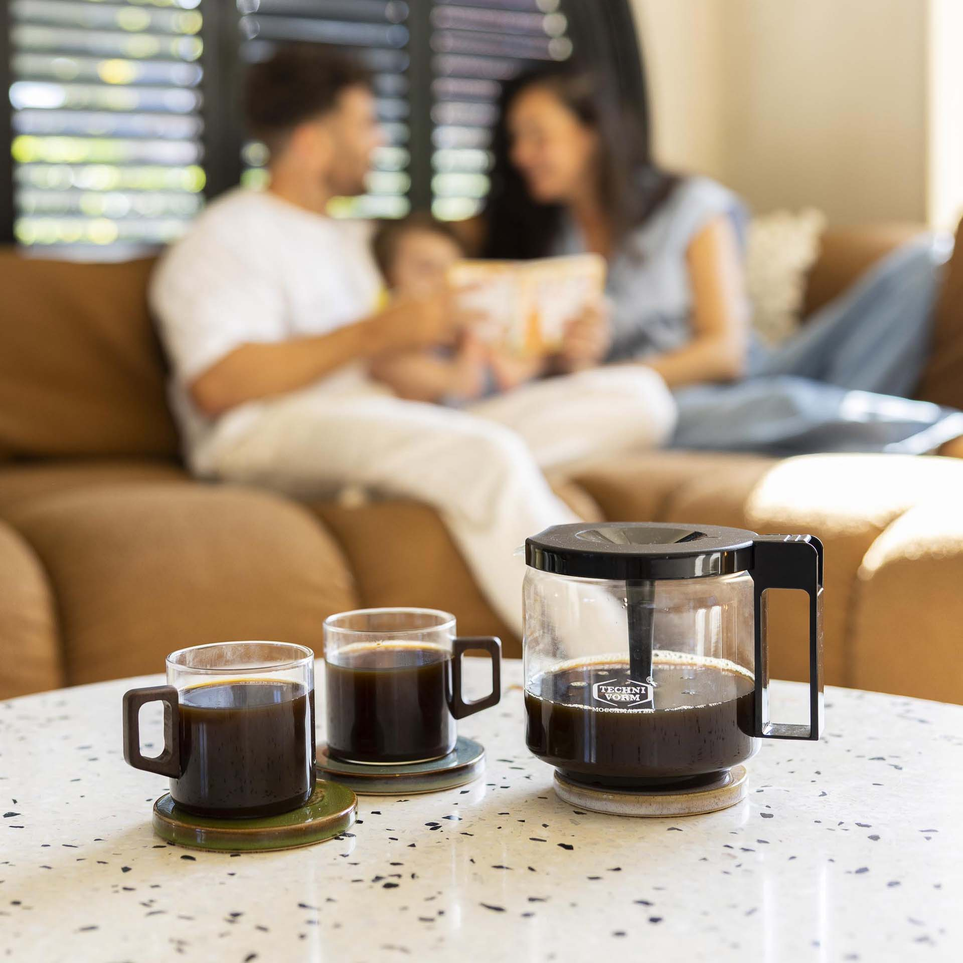 Une cafetière avec deux tasses de café sur une table, famille en arrière-plan.