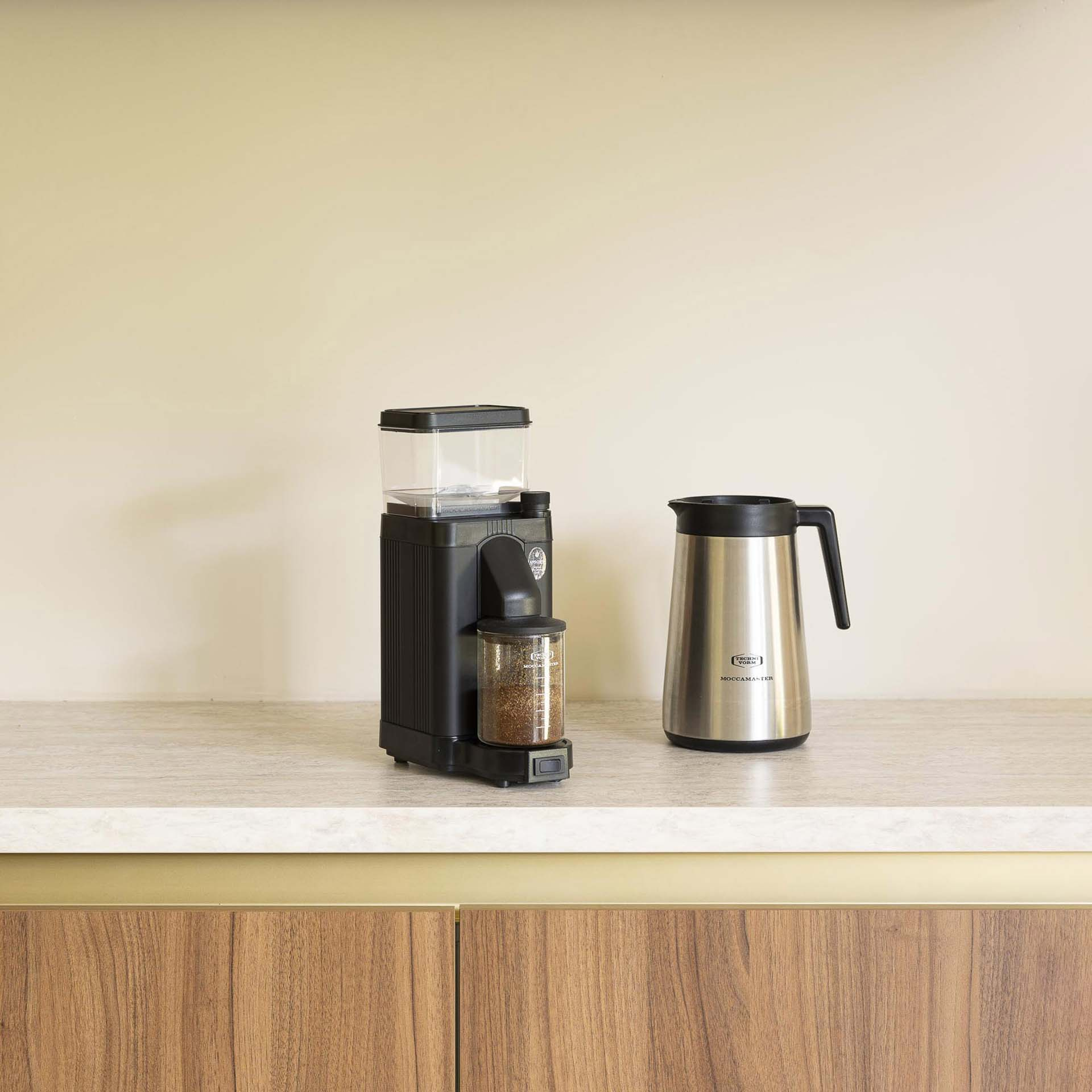 Ein schwarzer Kaffeemühle und eine silberne Thermoskanne stehen auf einer Arbeitsplatte.