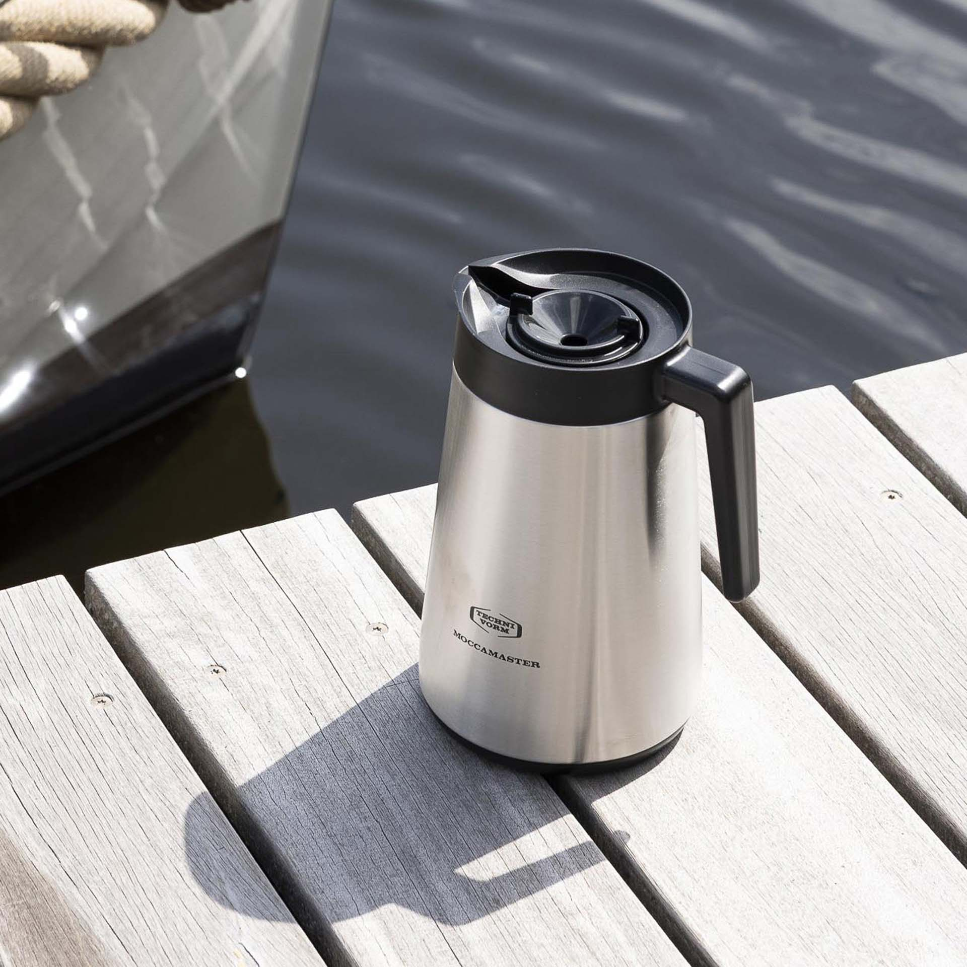 Silberne Thermoskanne mit schwarzem Griff und Deckel auf Holzplanken am Wasser.