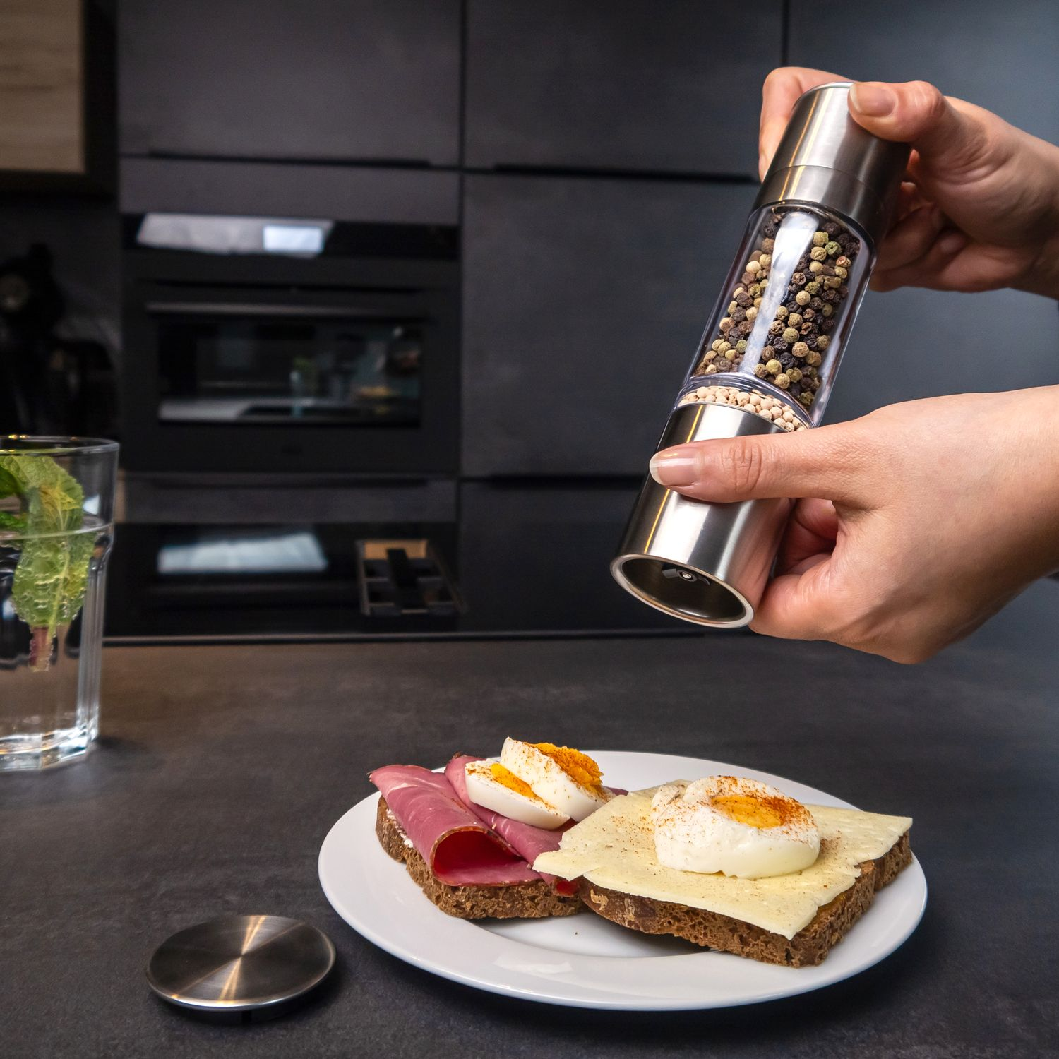 Eine Hand mahlt Pfeffer über ein Sandwich mit Käse, Schinken und Ei in einer Küche.