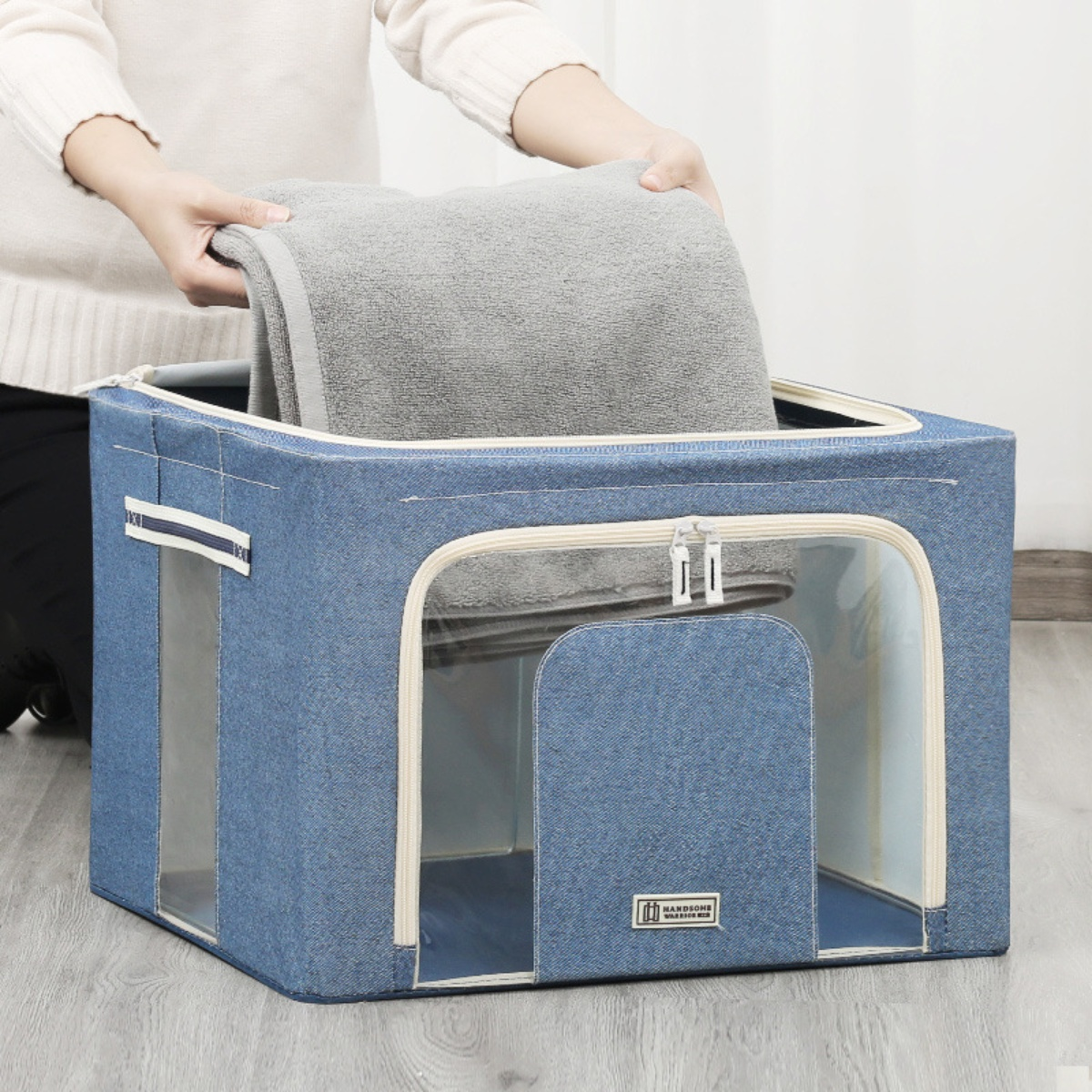 Eine Person legt ein graues Handtuch in eine blaue Stoffbox mit transparenten Fenstern.