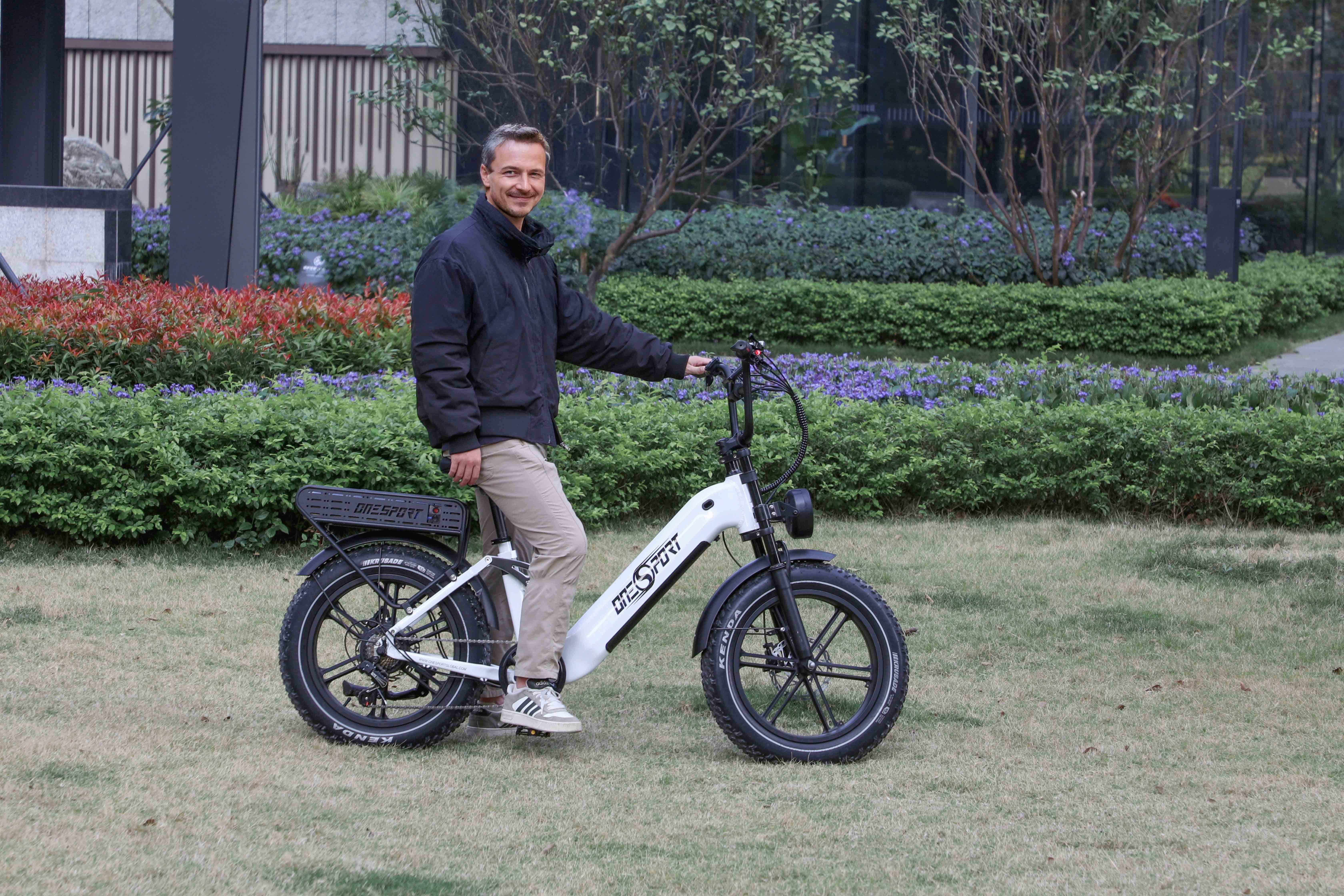 Mann in schwarzer Jacke steht neben weißem E-Bike im Gras mit Büschen.