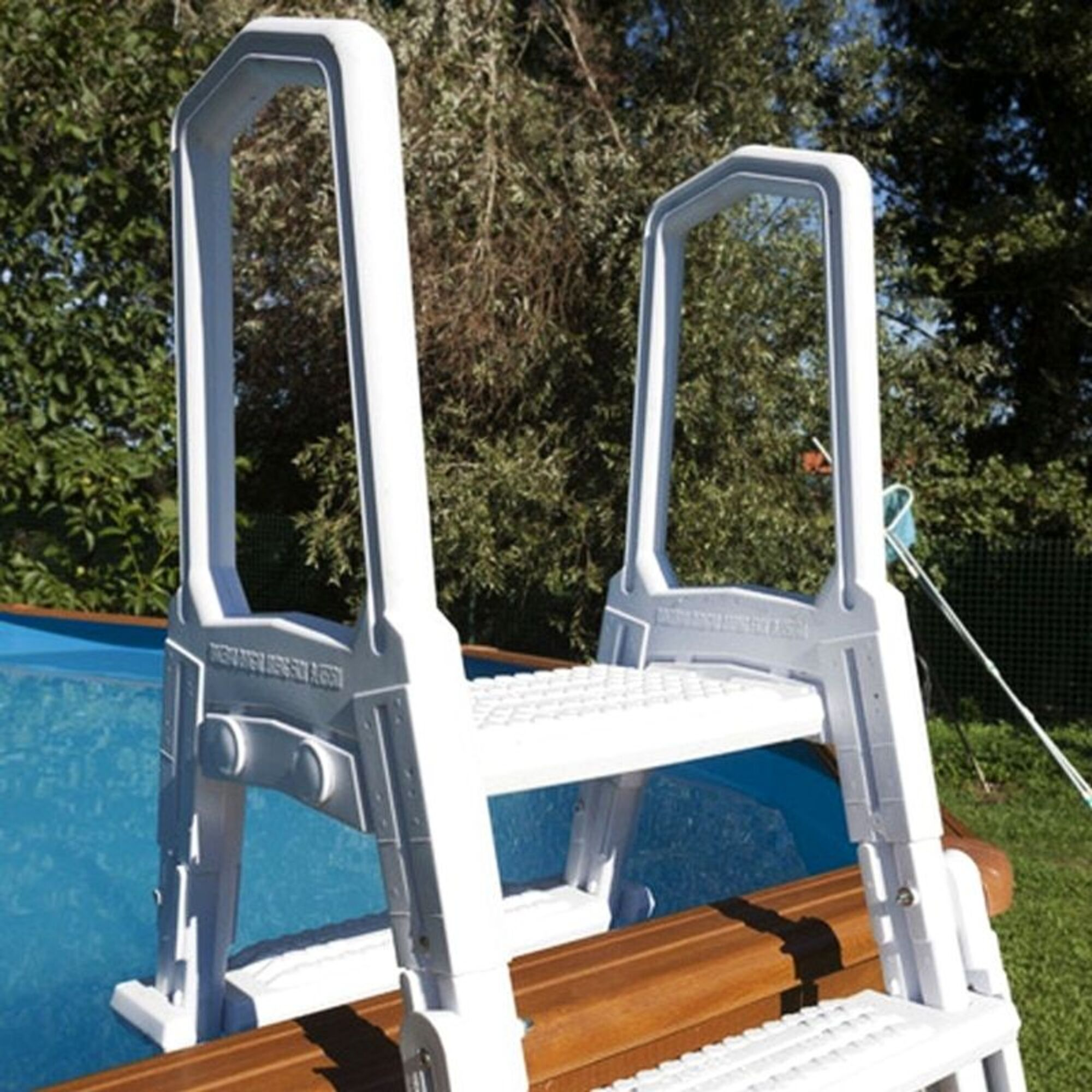 Una escalera de piscina blanca está parcialmente en agua azul. Árboles al fondo.
