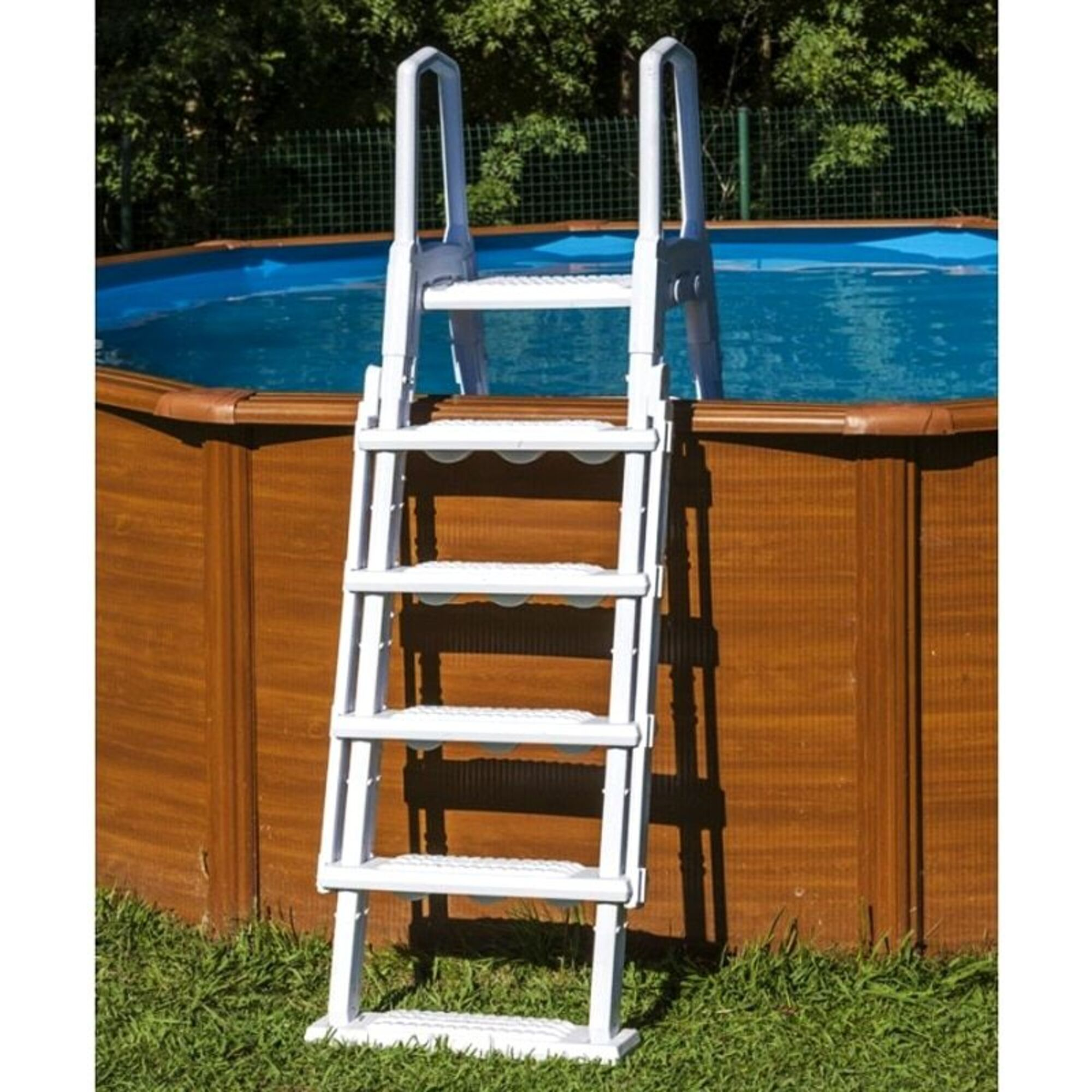 Una escalera de piscina blanca está sobre césped junto a una piscina de madera con agua azul.