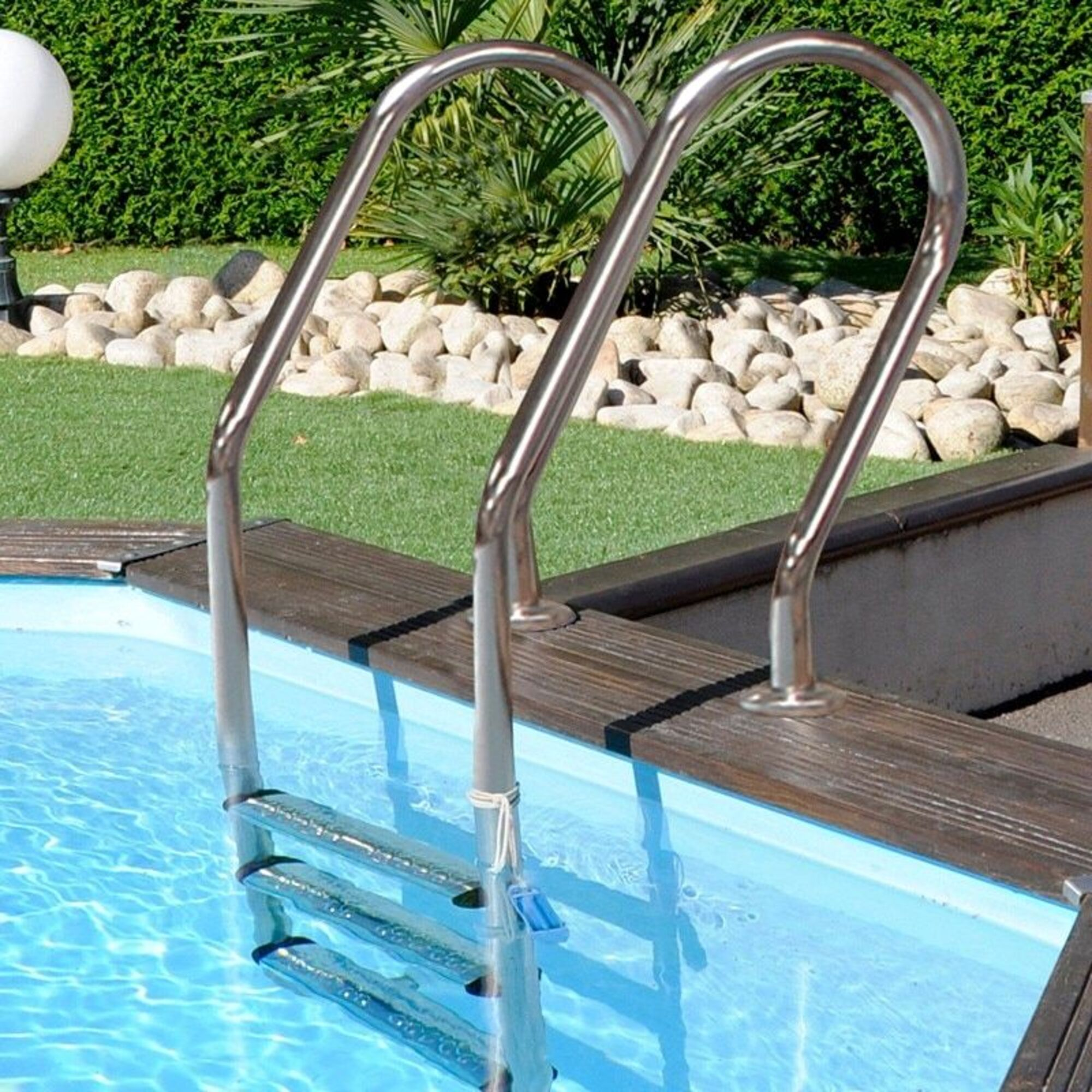 Una escalera de piscina de acero inoxidable sale del agua azul. Cubierta de madera.