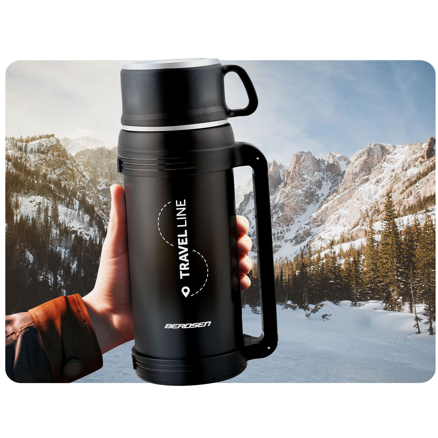 Dłoń trzyma czarny termos na tle zaśnieżonego krajobrazu górskiego z drzewami.