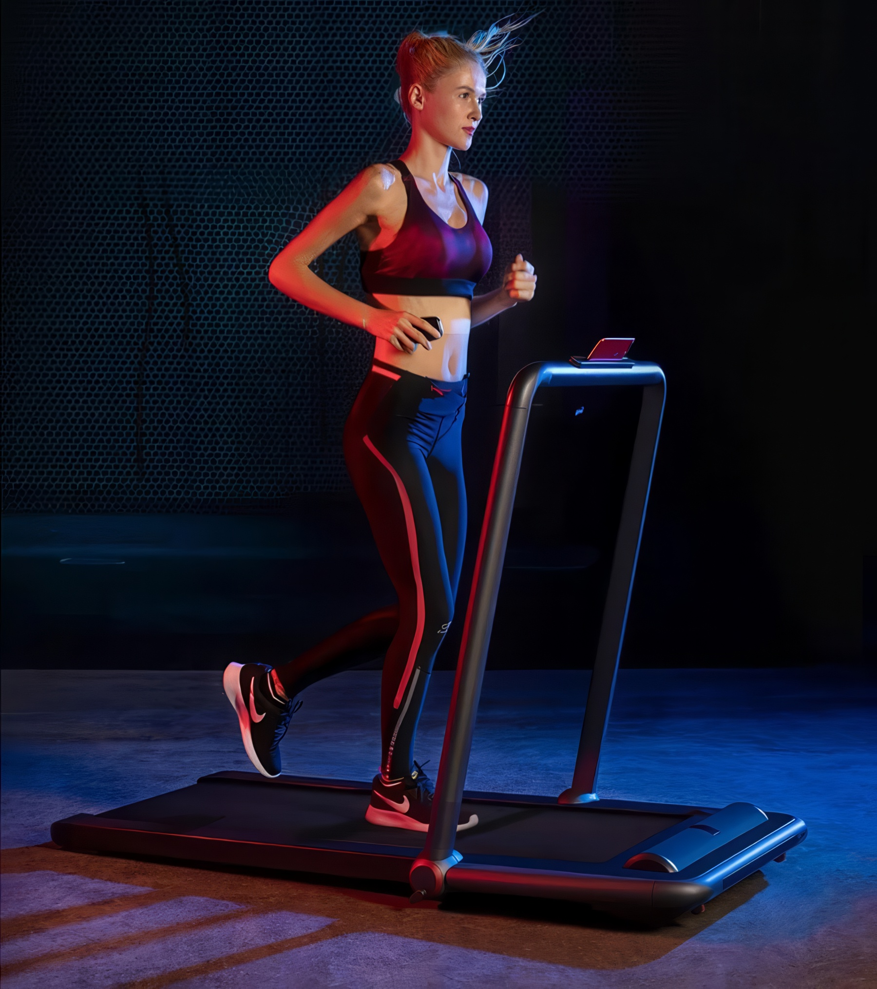Kobieta biegnie na bieżni w ciemnym pomieszczeniu z niebieskimi i czerwonymi światłami.