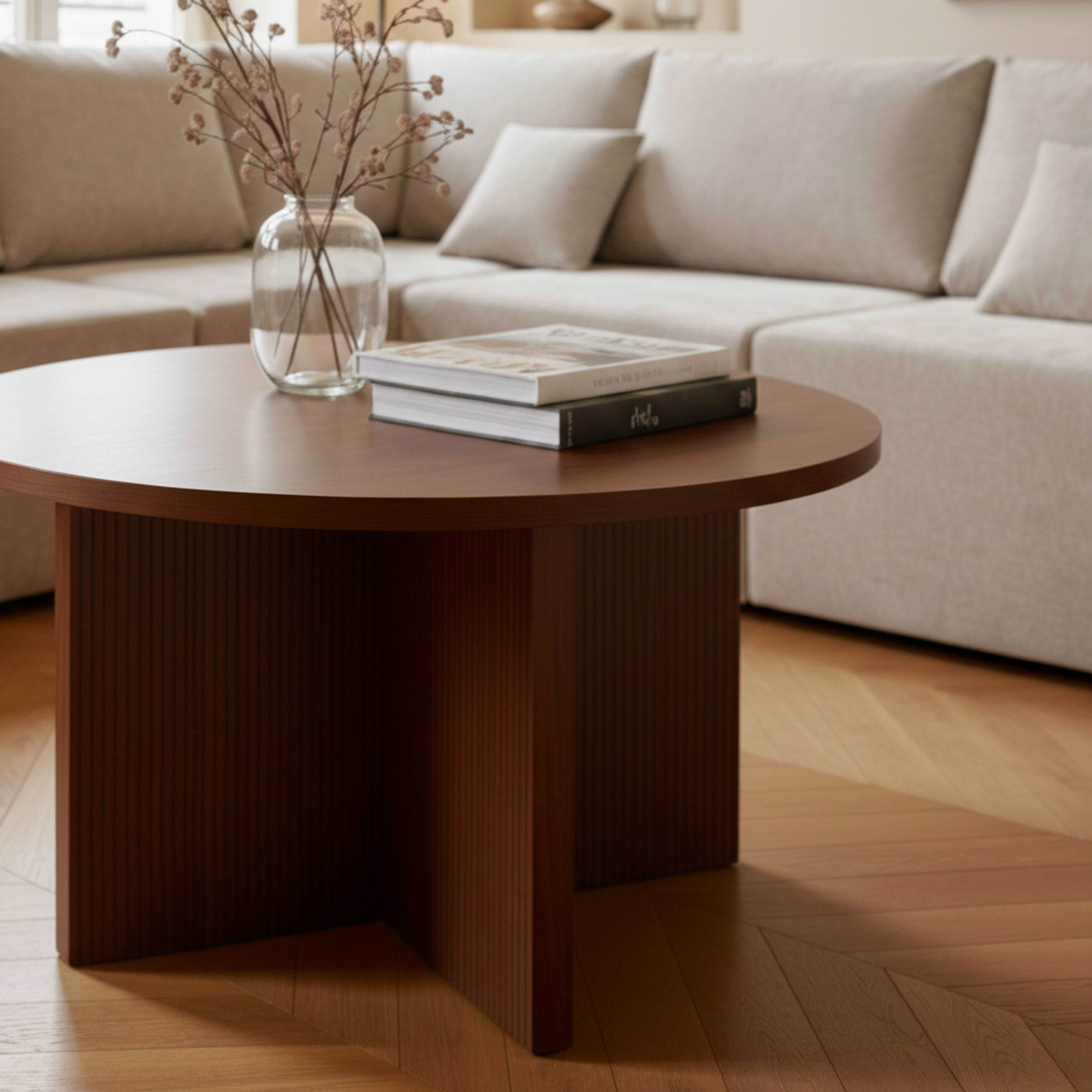 Ein runder Holztisch mit Büchern und einer Vase mit Zweigen steht vor einem Sofa.