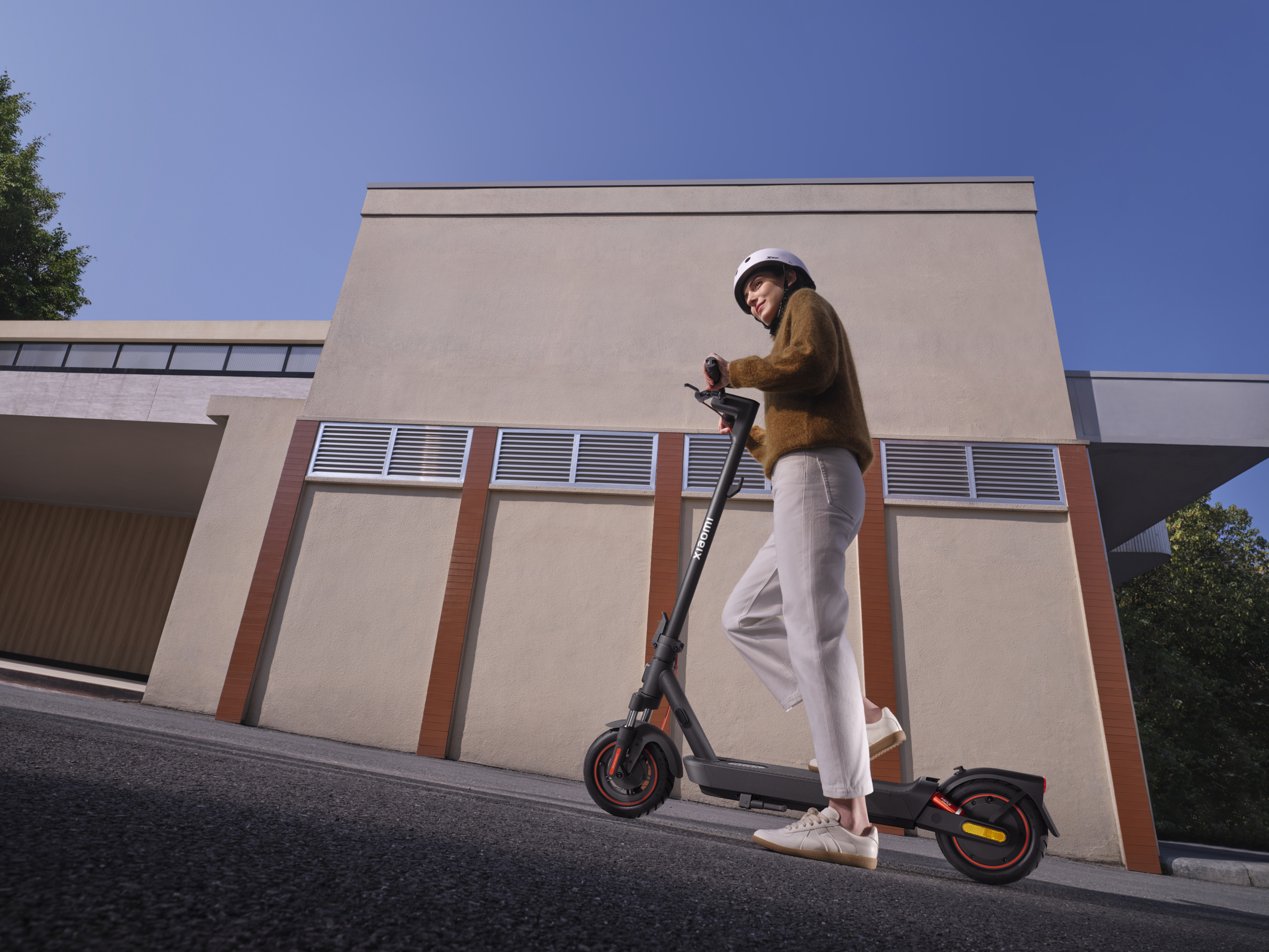 Kobieta w kasku jedzie na hulajnodze elektrycznej przed budynkiem przy błękitnym niebie.