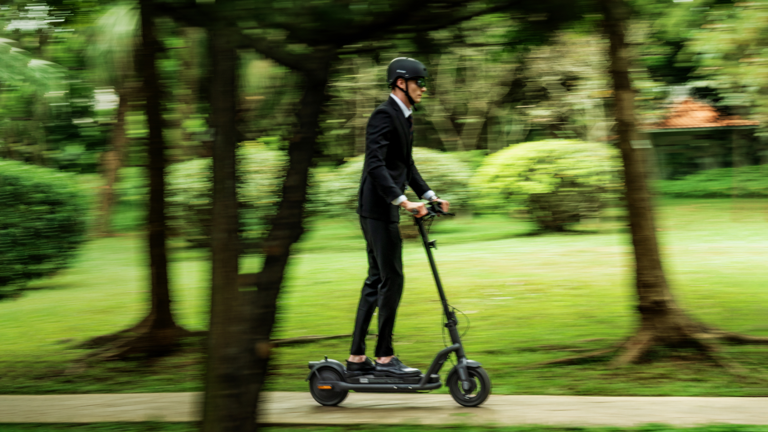 Mann im Anzug fährt E-Scooter durch grünen Park mit Bäumen.