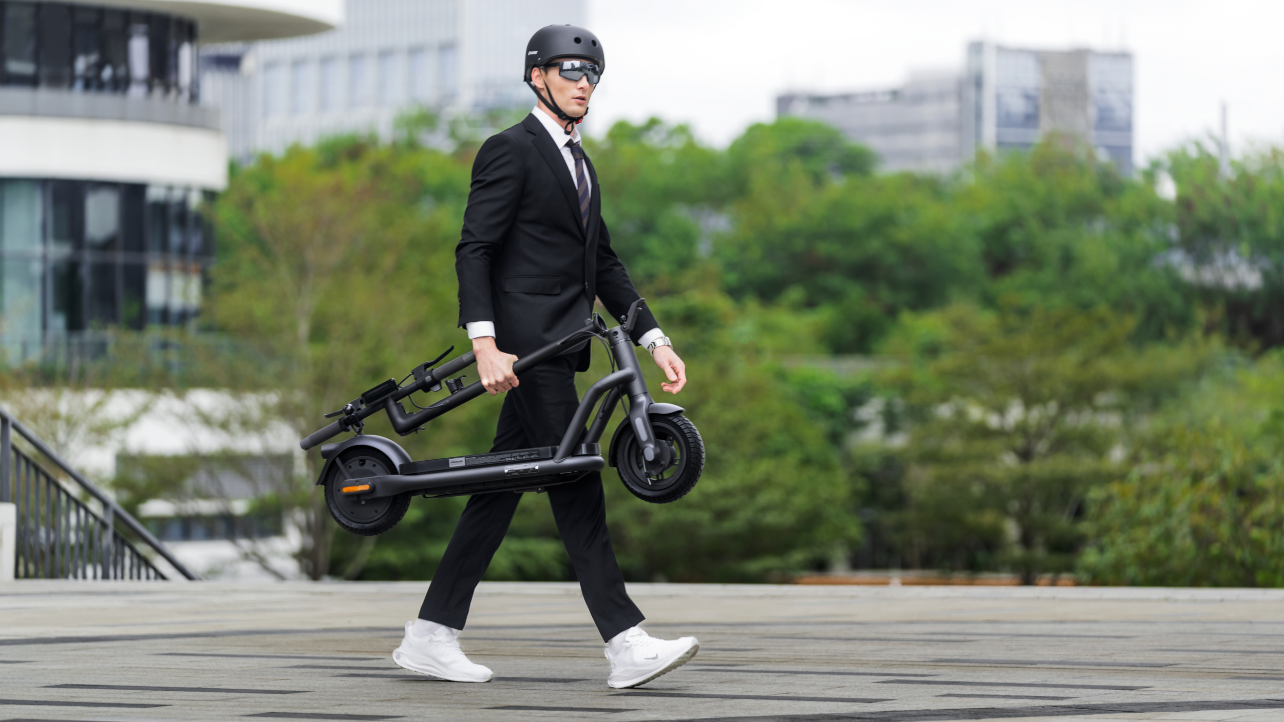 Ein Mann im schwarzen Anzug trägt einen zusammengeklappten Roller. Er trägt einen Helm und Sonnenbrille.