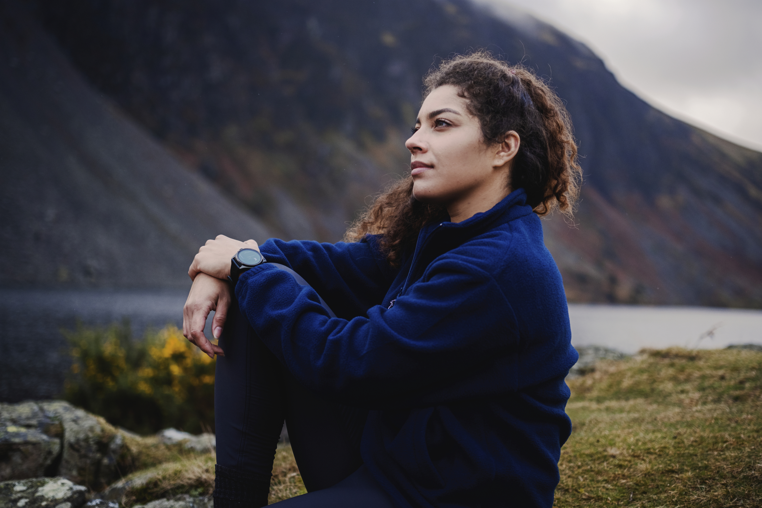 Eine junge Frau sitzt auf einem Felsen mit Blick auf einen See und Berge.