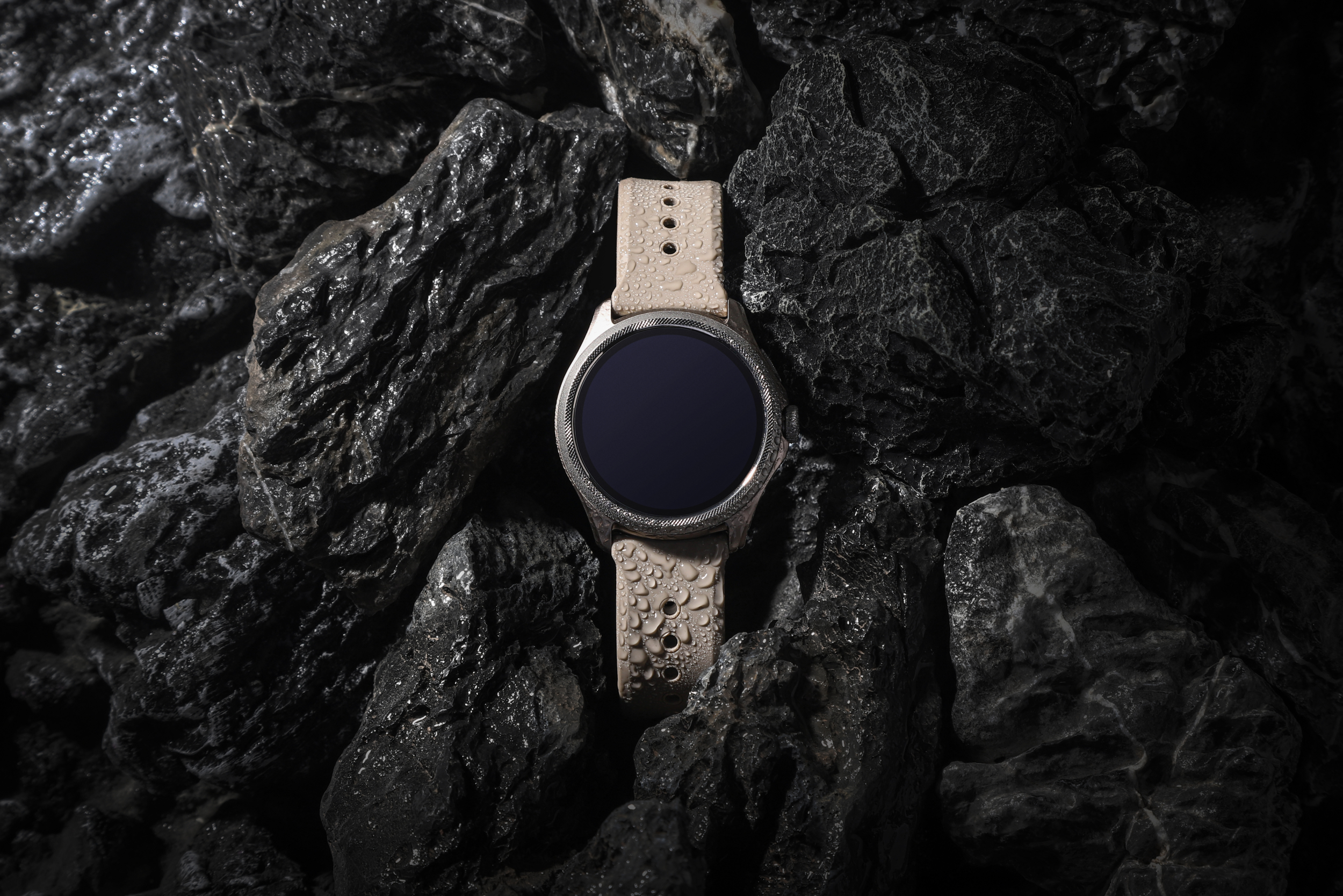 Jasny smartwatch z beżowym paskiem spoczywa na ciemnych, mokrych skałach.