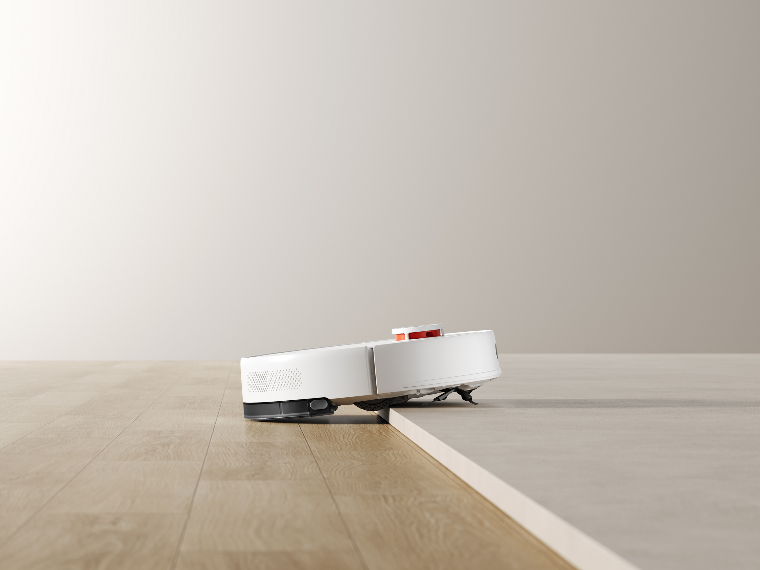 Kırmızı vurgulu beyaz bir robot süpürge ahşap zeminde bir kenarın üzerinden geçiyor.