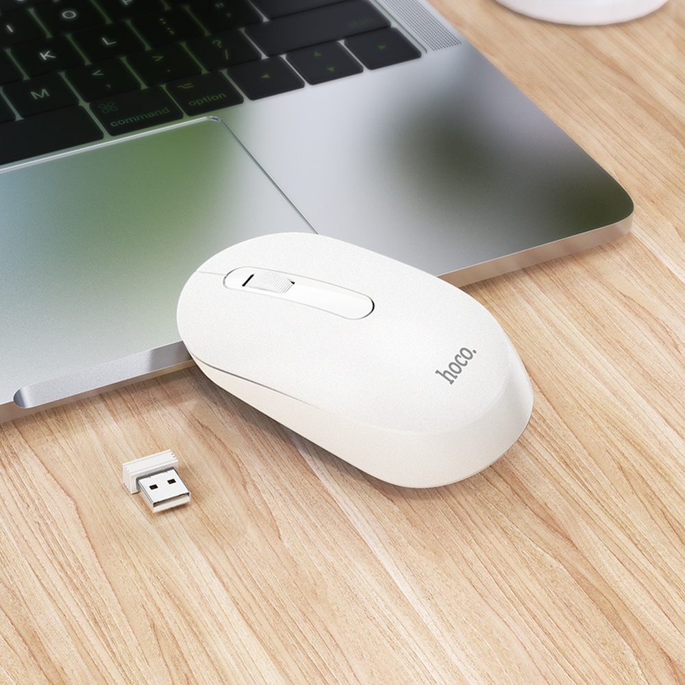 Un ratón de ordenador blanco y un dongle USB descansan sobre un escritorio de madera junto a un portátil.