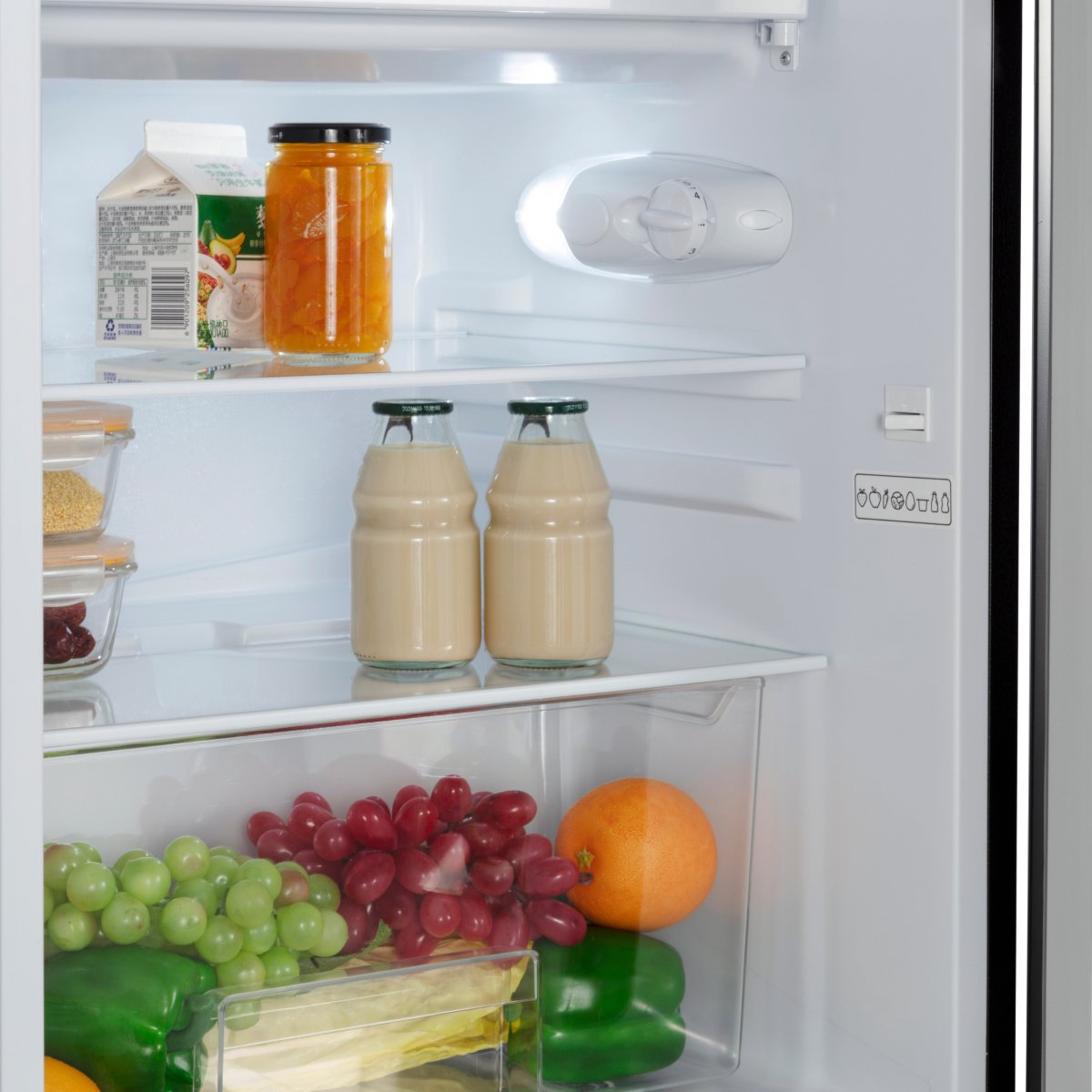 Réfrigérateur ouvert avec fruits, boissons et bocaux sur les étagères. Raisins verts et rouges, orange, poivrons.
